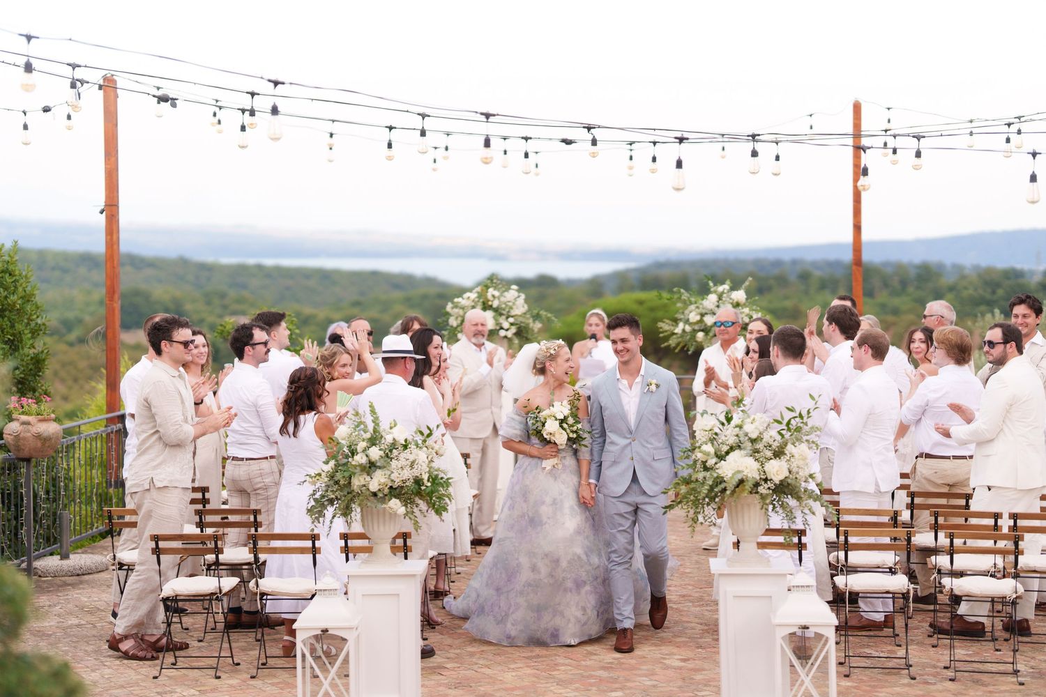 Outdoor wedding ceremony on a scenic terrace with string lights, white floral arrangements, and guests in elegant attire.