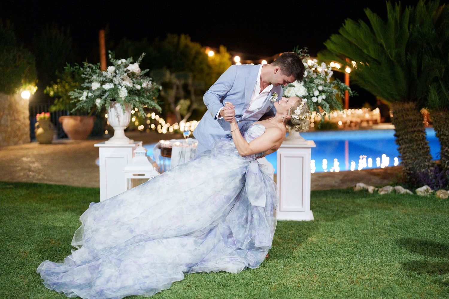 Bride and groom share romantic dip kiss at outdoor evening wedding reception near pool with floral arrangements.