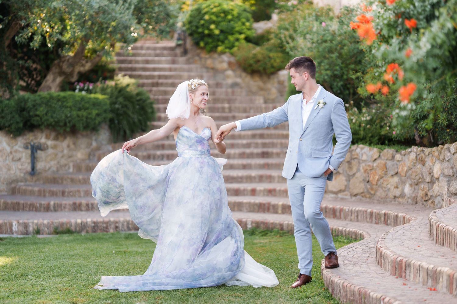 Bride in blue watercolor gown dances with groom in gray suit outdoors on lush garden steps.