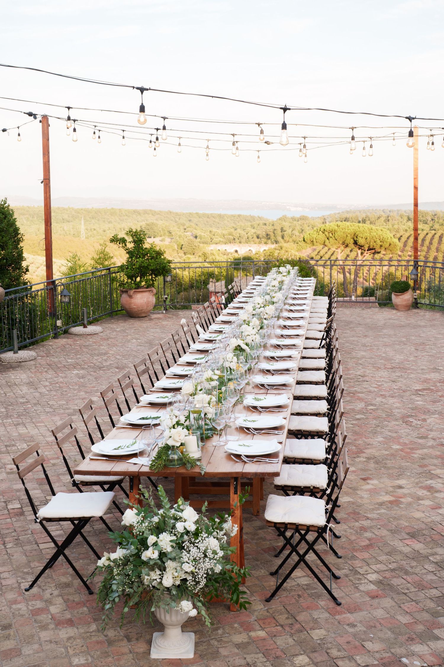 Elegant outdoor wedding reception with long wooden table, string lights, white floral arrangements, and vineyard views.