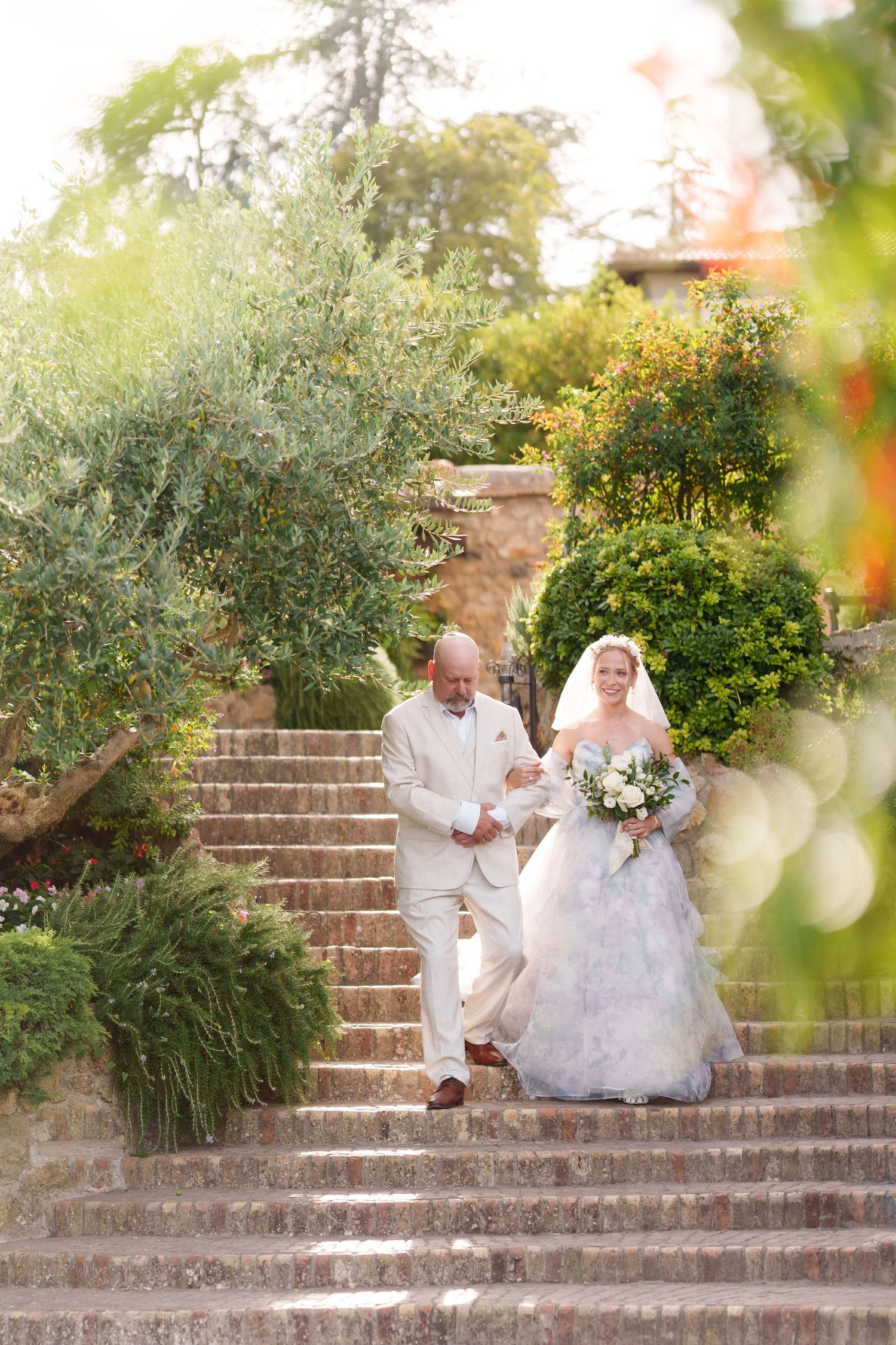 Bride in white gown and groom in tan suit descend stone steps surrounded by lush greenery at outdoor wedding.