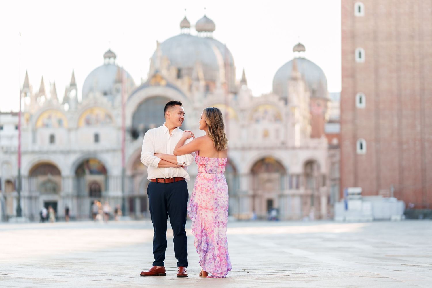 Couple holding hands in St. Mark's Square, Venice, with the iconic Basilica di San Marco glowing in golden light behind them.