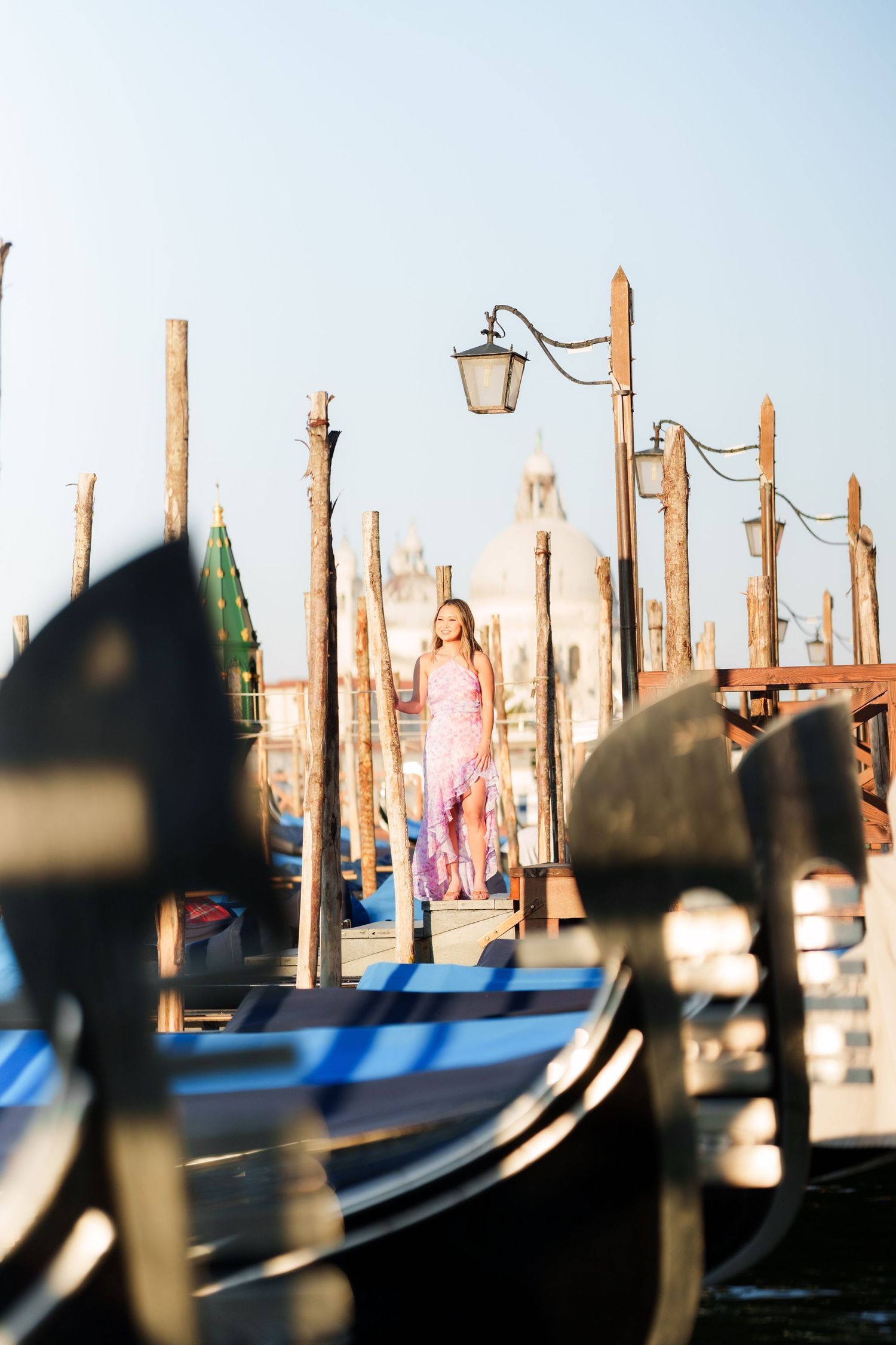 Woman in pink dress standing on Venice gondola dock with wooden poles and street lamp at golden hour.