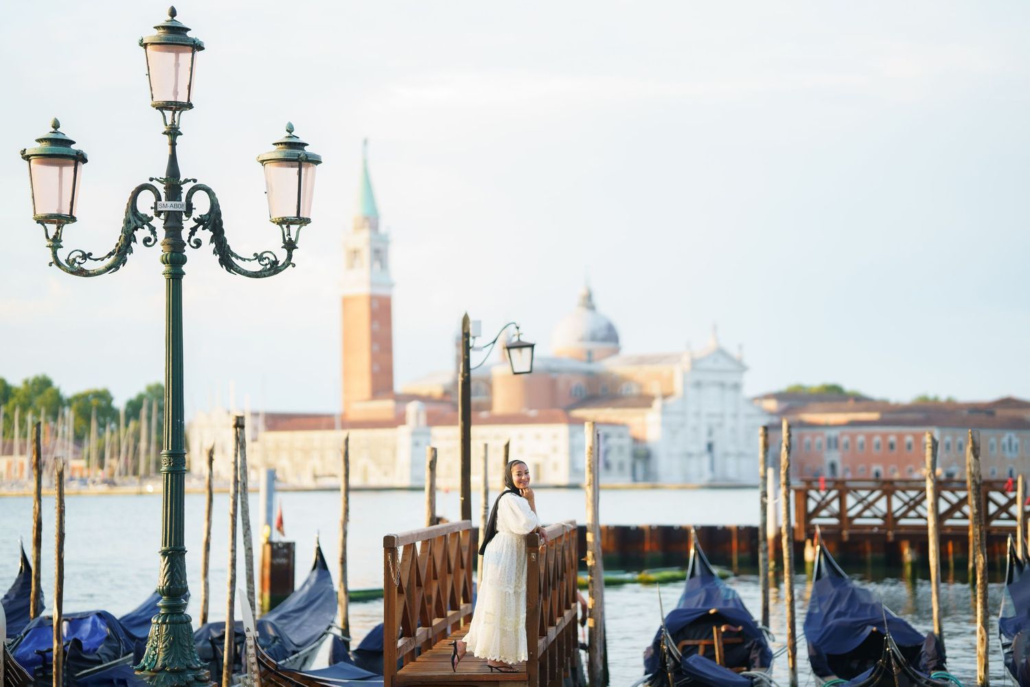 Woman in white dress standing on Venice dock with gondolas and San Giorgio Maggiore church in background.