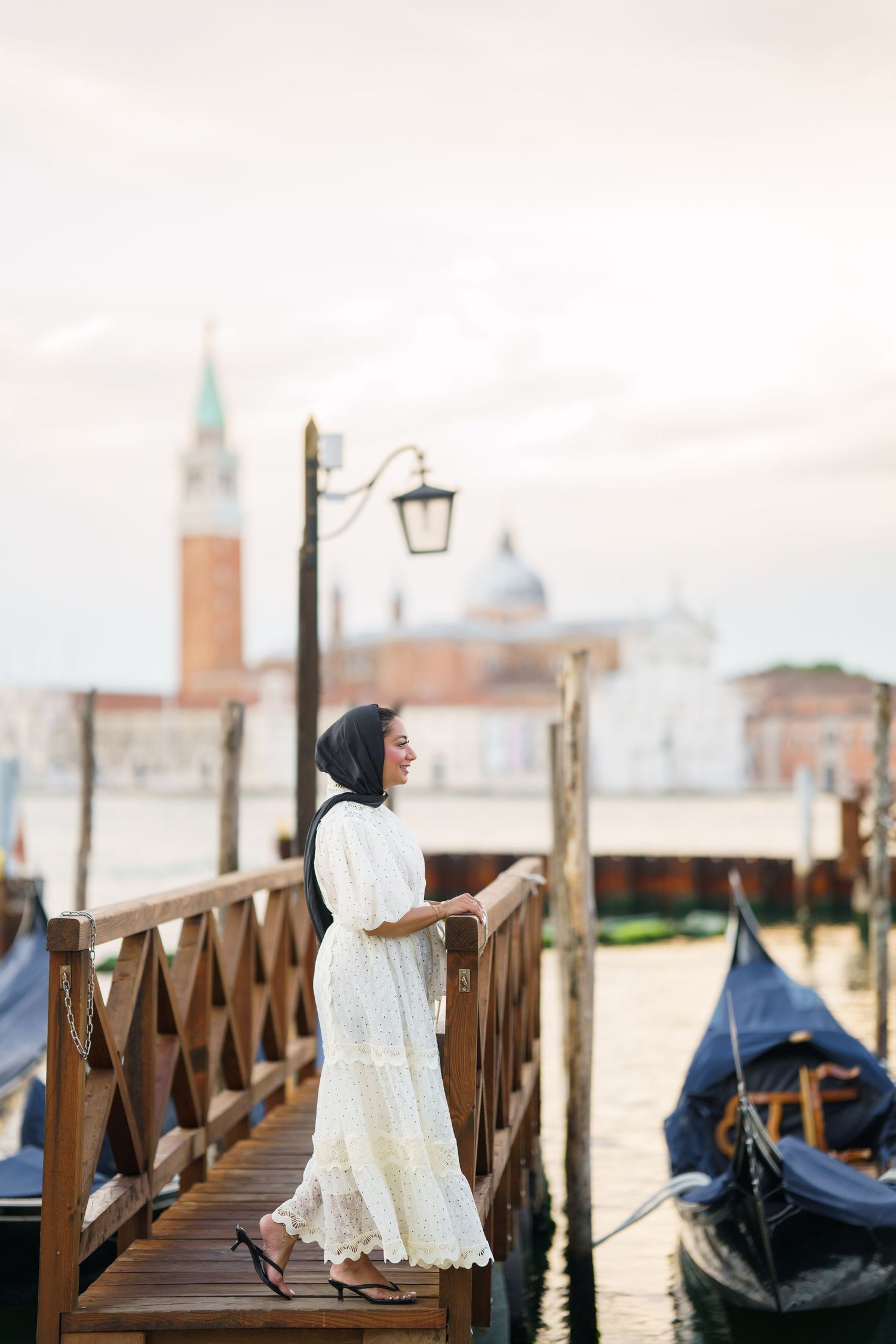 Woman in white dress and black hijab standing on a wooden dock in Venice, Italy with gondolas and St. Mark's.