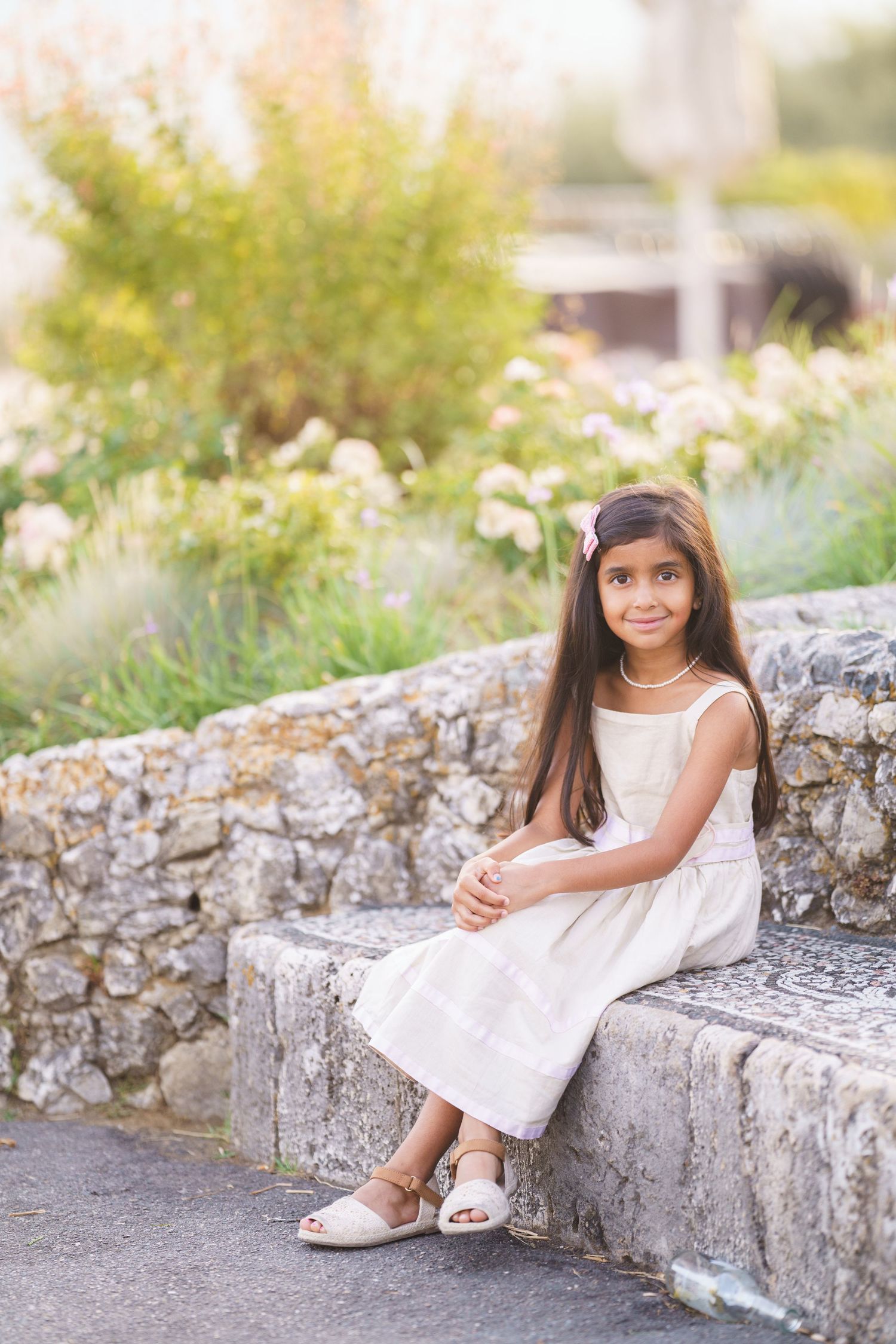 Young girl in white dress sitting on stone steps surrounded by lush garden greenery.