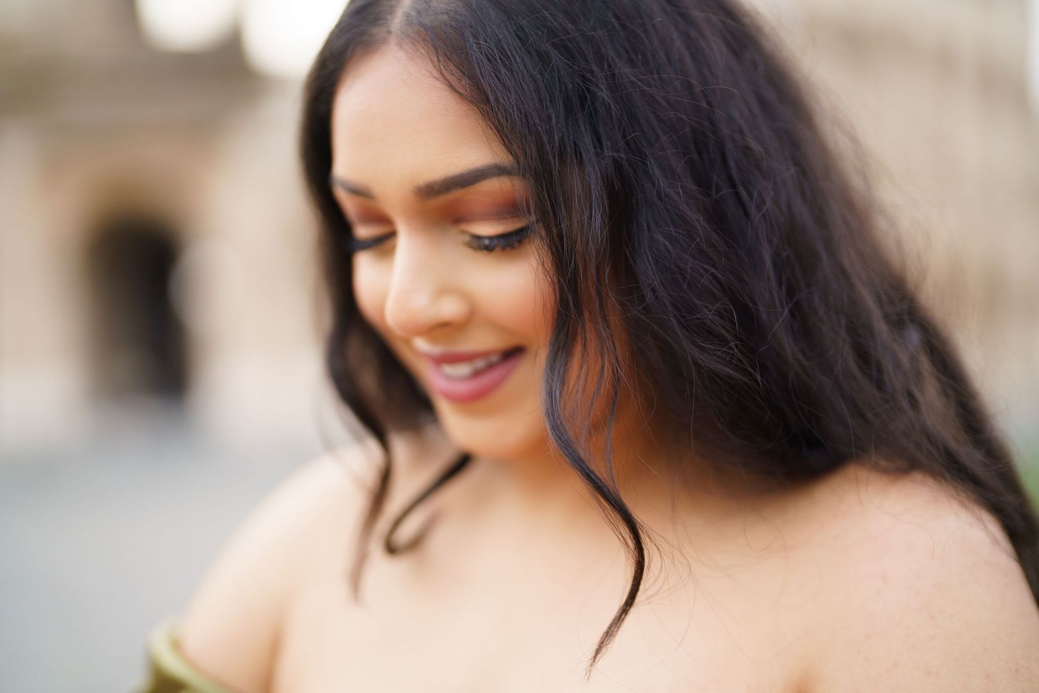A young South Asian woman with long dark hair smiles softly outdoors, wearing an off-shoulder top in natural light.