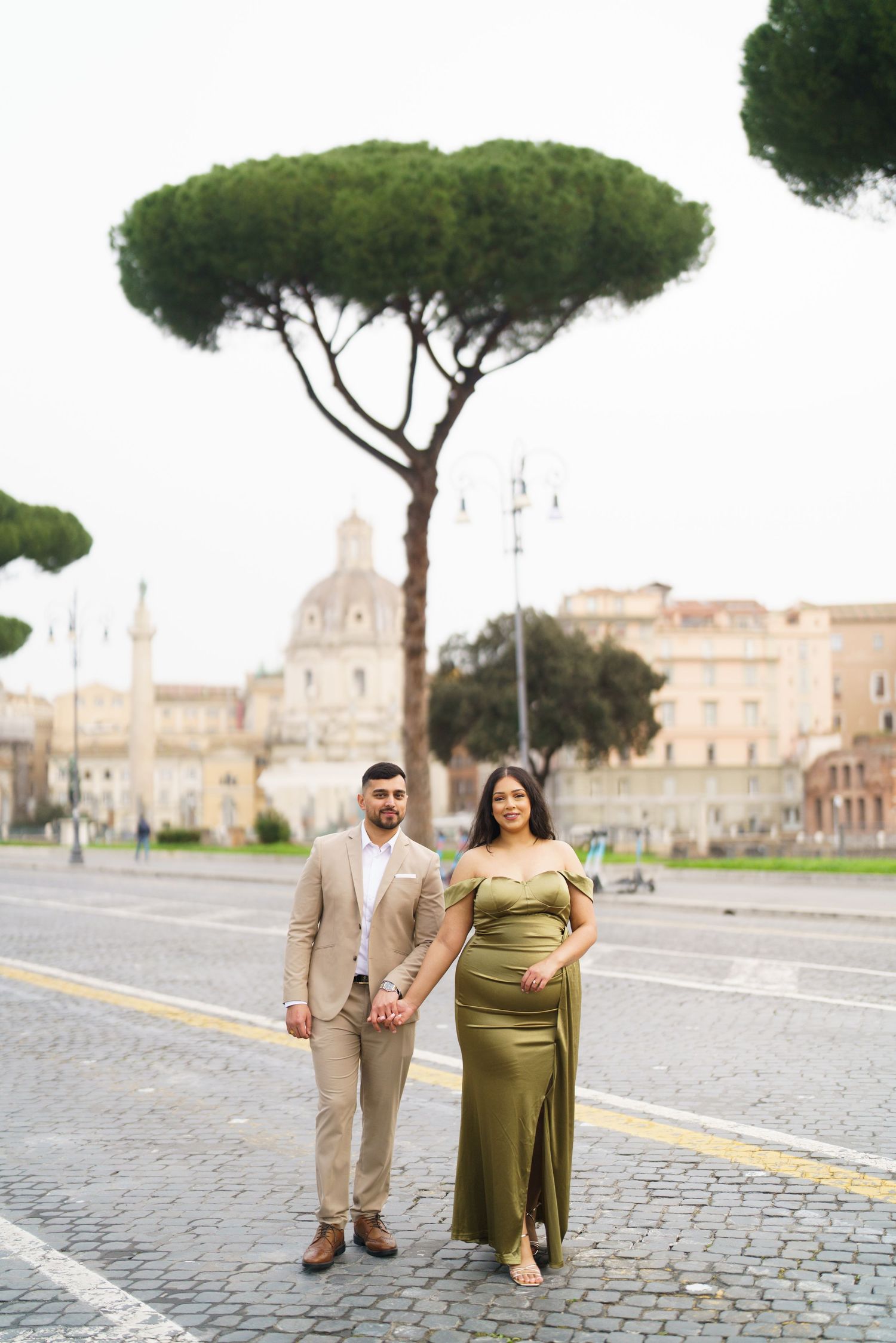 Couple posing on a cobblestone street in Rome, Italy, with iconic umbrella pine trees and ancient ruins in the background.