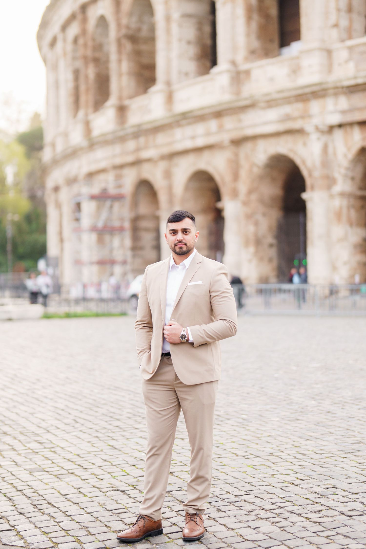 A well-dressed man in a beige suit stands confidently in front of the Roman Colosseum on cobblestone pavement.