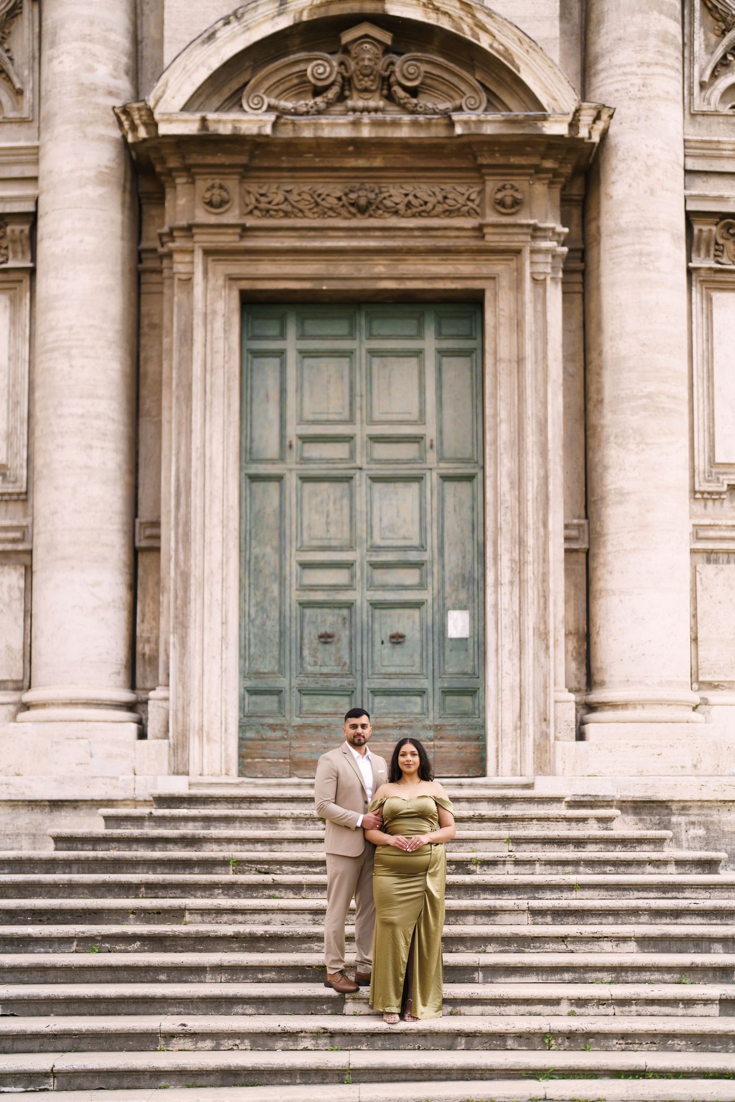 Elegant couple posing on stone steps before a grand classical building with ornate green doors.