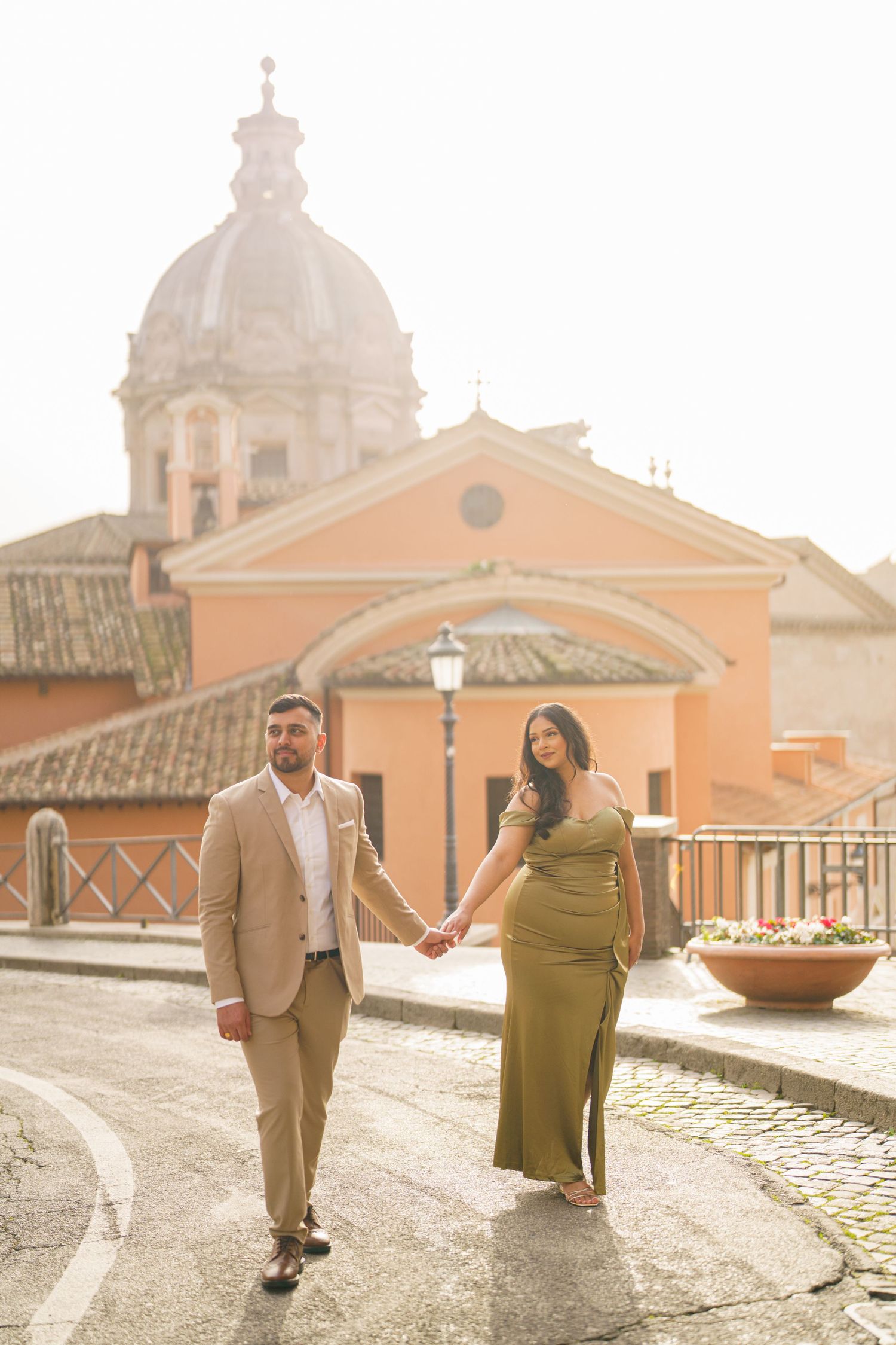 Couple holding hands walking on cobblestone street in Rome with historic church dome and warm golden light in background.