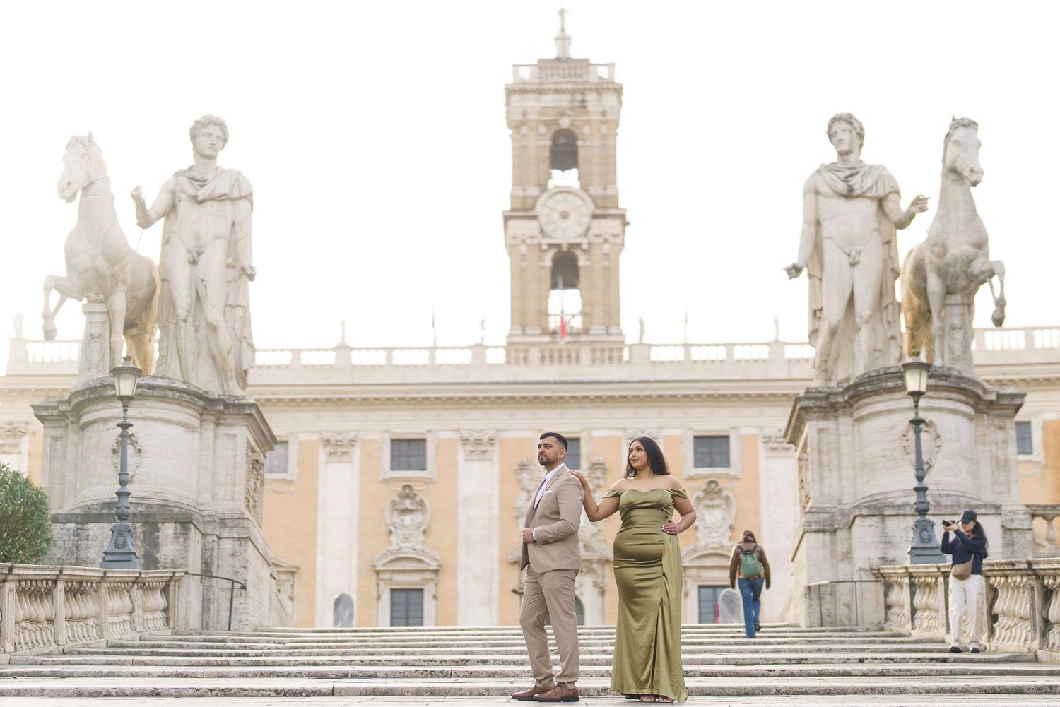 Couple posing on the steps of Piazza del Campidoglio in Rome, Italy, with classical statues and a historic building behind them.