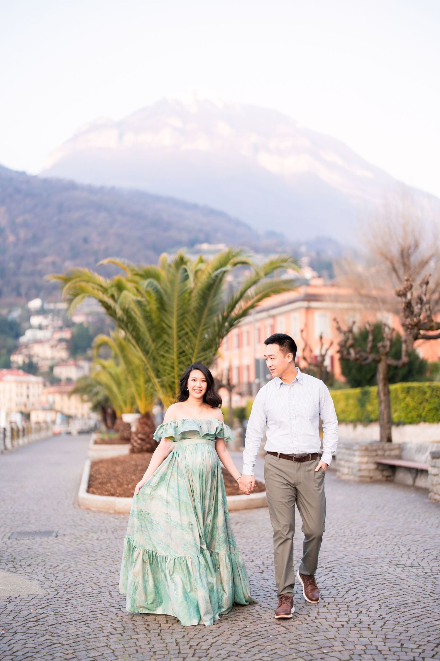 Couple holding hands walking on cobblestone path with palm trees and mountain backdrop at golden hour.