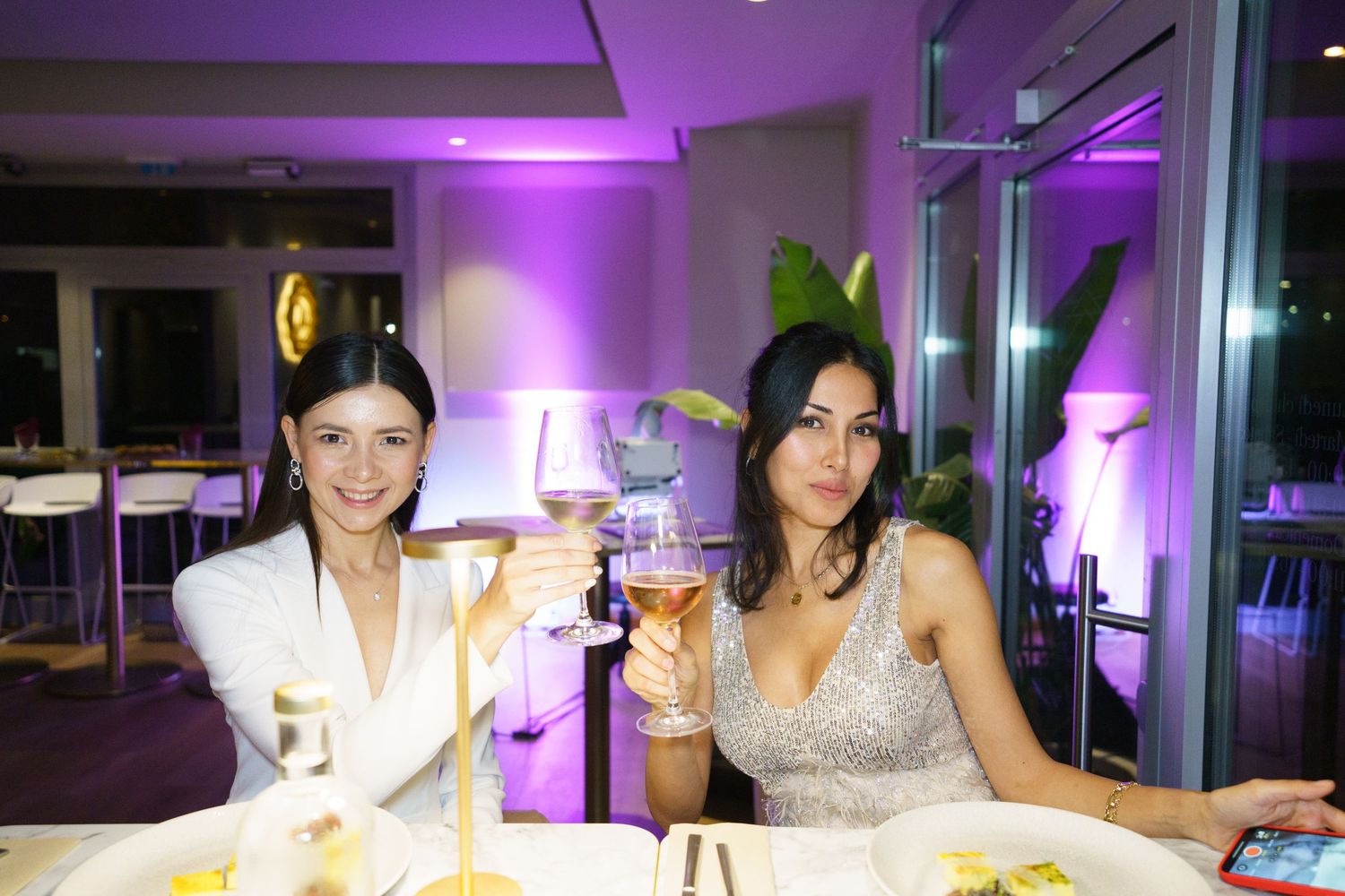 Two women toasting with wine glasses at an elegant restaurant with purple ambient lighting.