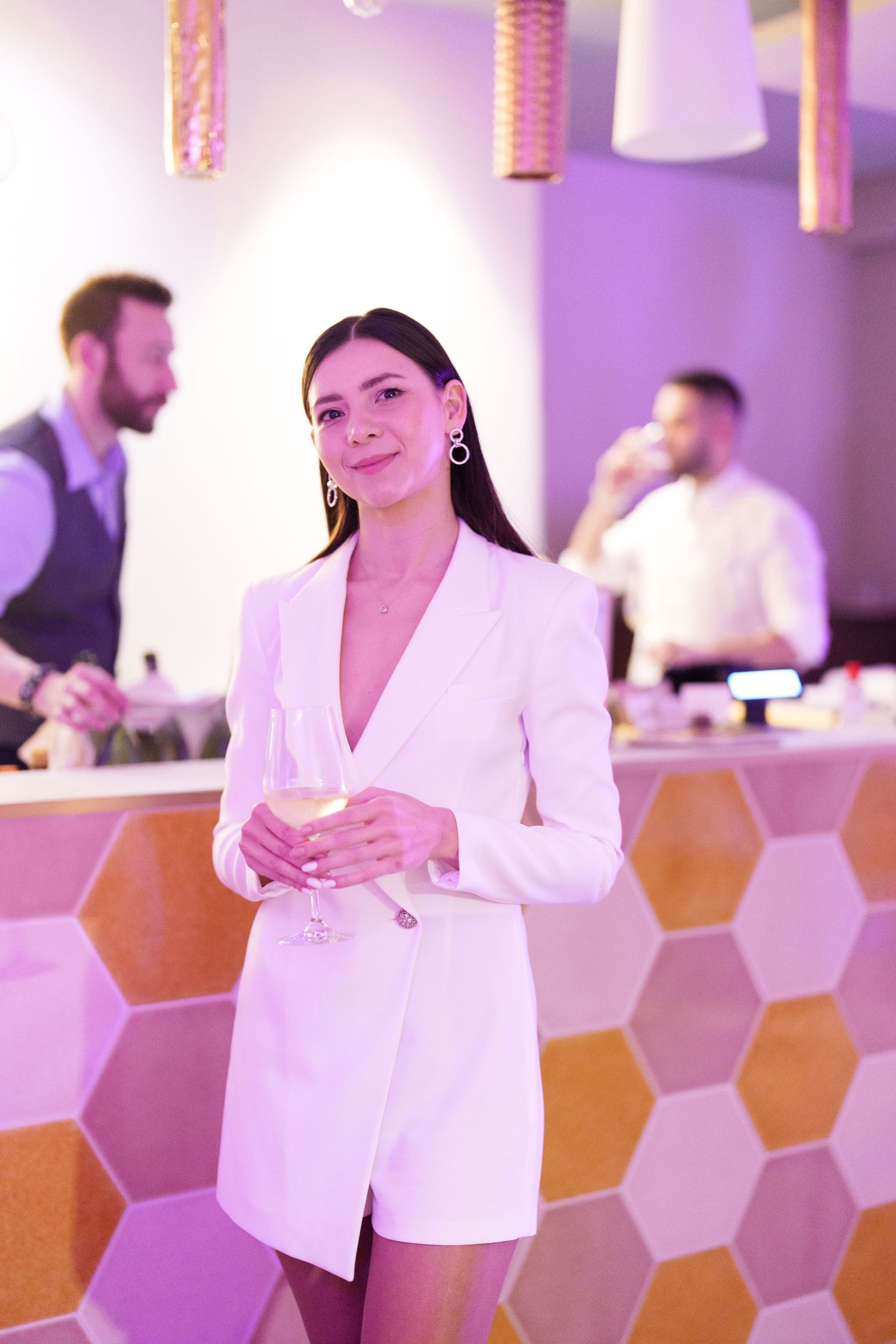 Elegant woman in white blazer dress holding champagne glass at a stylish bar with hexagonal tile decor.