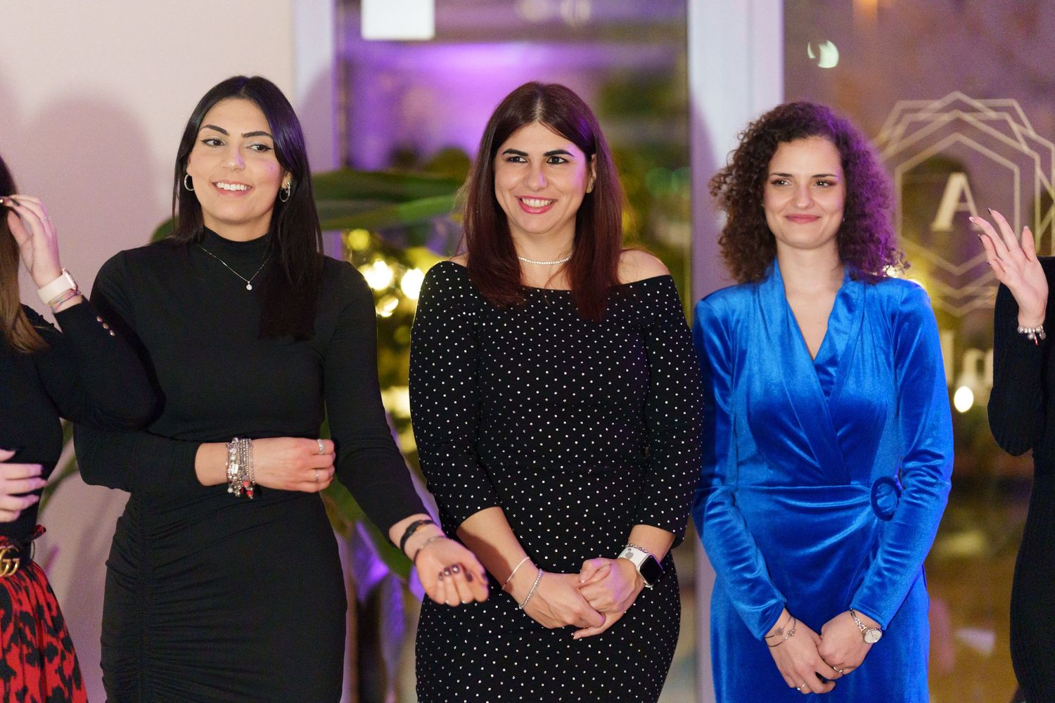 Three women smiling and posing together at an elegant indoor event, wearing black and blue dresses.