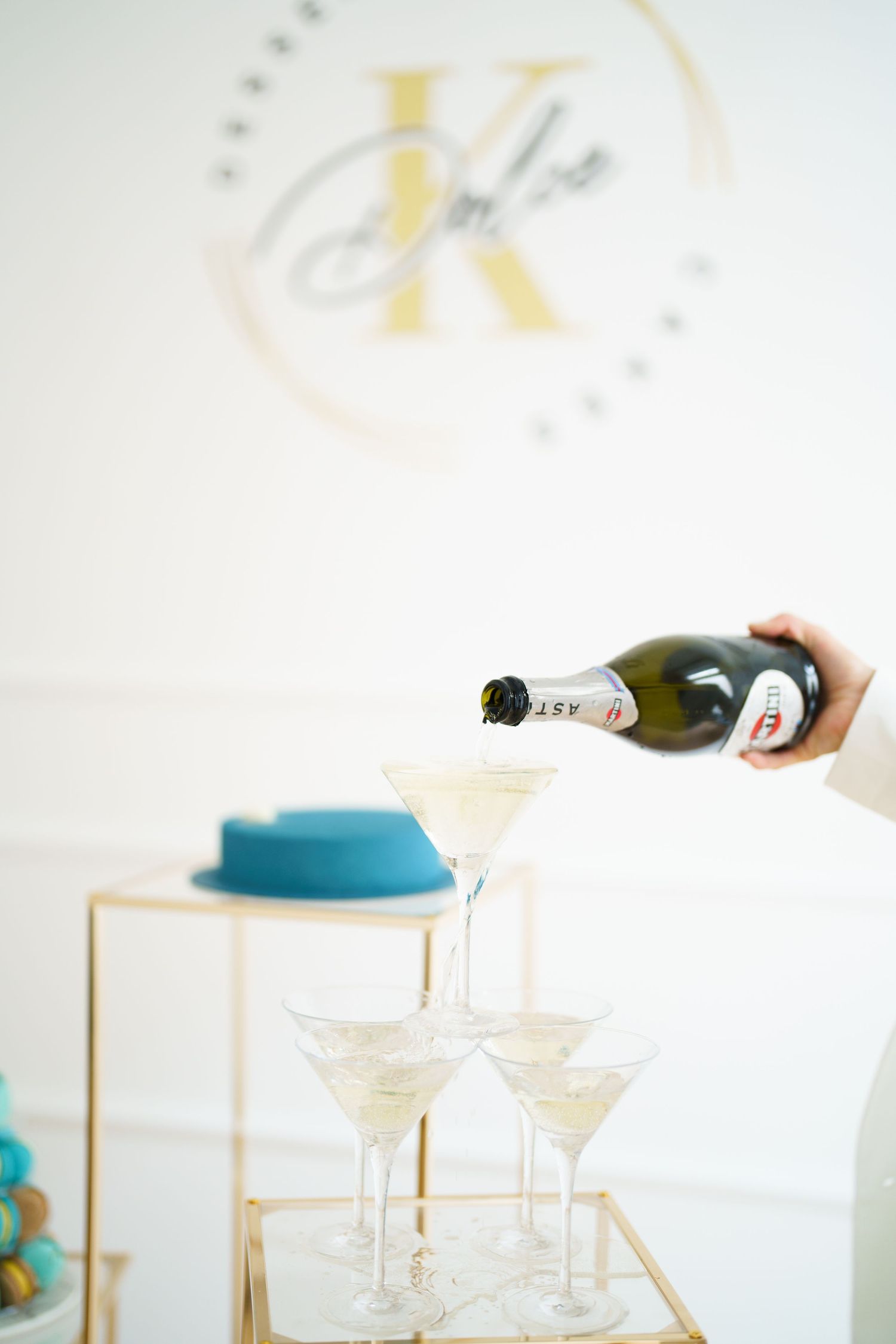 Champagne being poured into a pyramid of martini glasses at an elegant event with a teal cake and gold K logo backdrop.