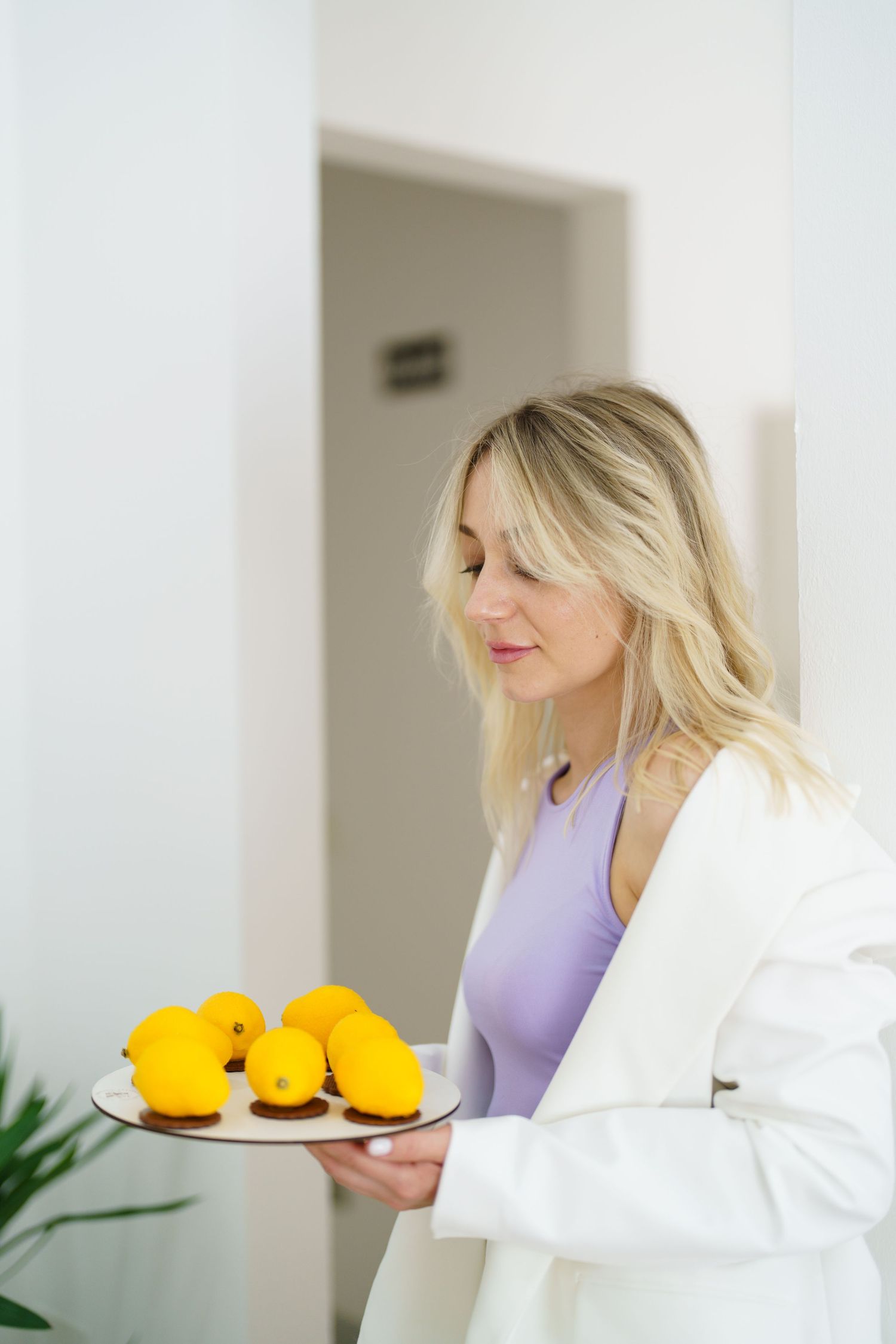 Blonde woman in white blazer holding a plate of fresh yellow lemons in a bright white room.