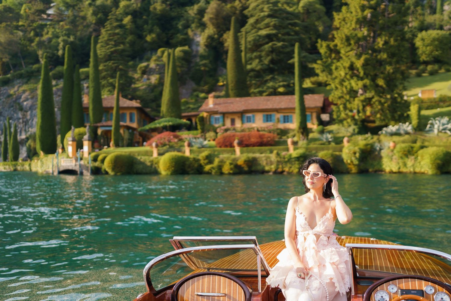 Woman in floral dress sitting on a wooden boat on turquoise lake waters with lush gardens and villa in background.
