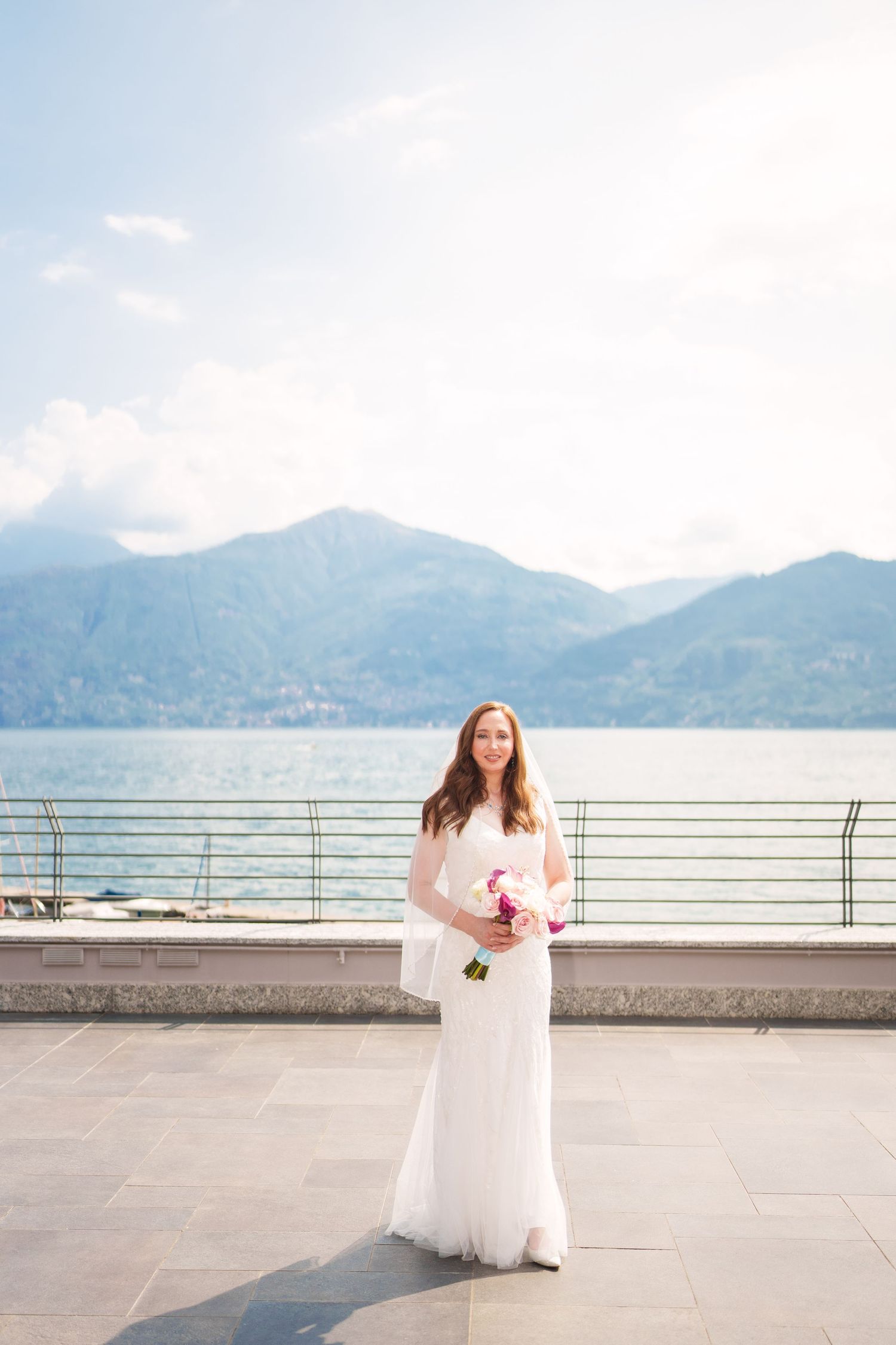 Bride in white gown holding pink bouquet stands on lakeside terrace with mountain backdrop on her wedding day como lake