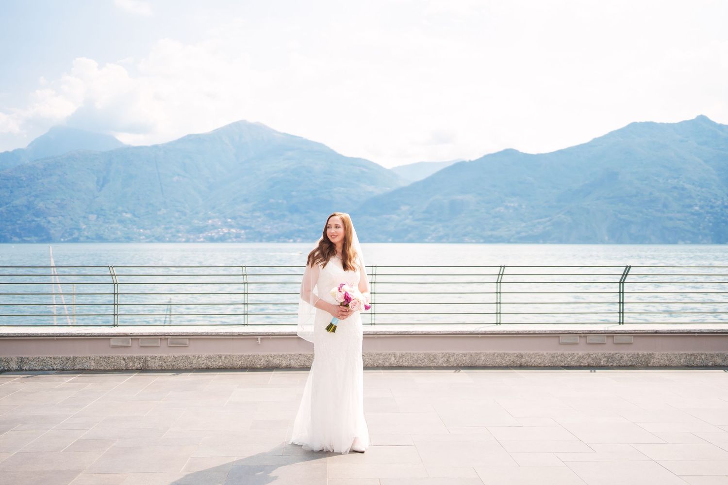 Bride in white gown holding bouquet on lakeside terrace with mountain backdrop on a bright day como lake