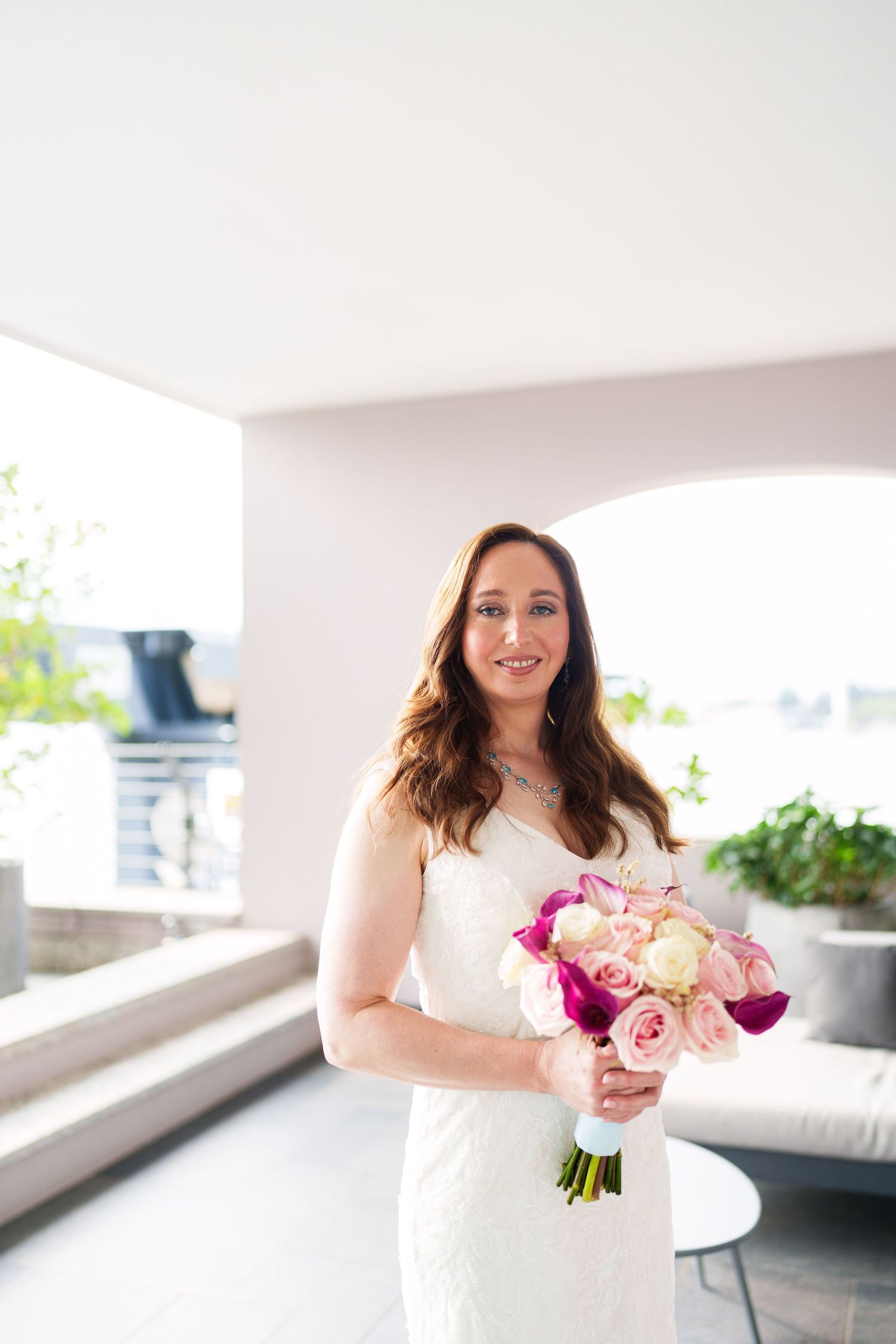 Bride in white lace gown holding pink and purple bouquet smiling on outdoor terrace.