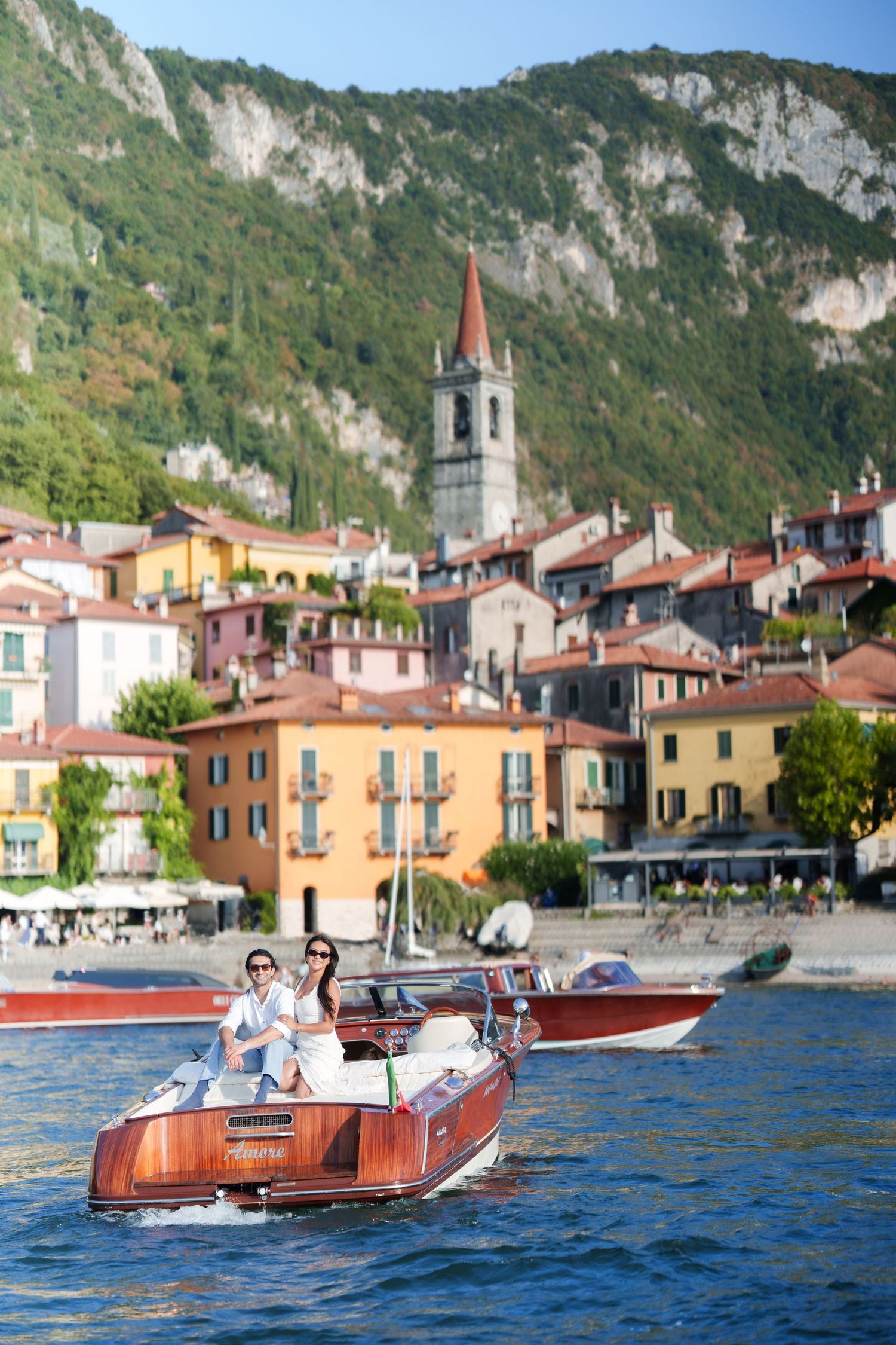 Couple riding a classic wooden motorboat on Lake Como with colorful Italian village and mountain backdrop.