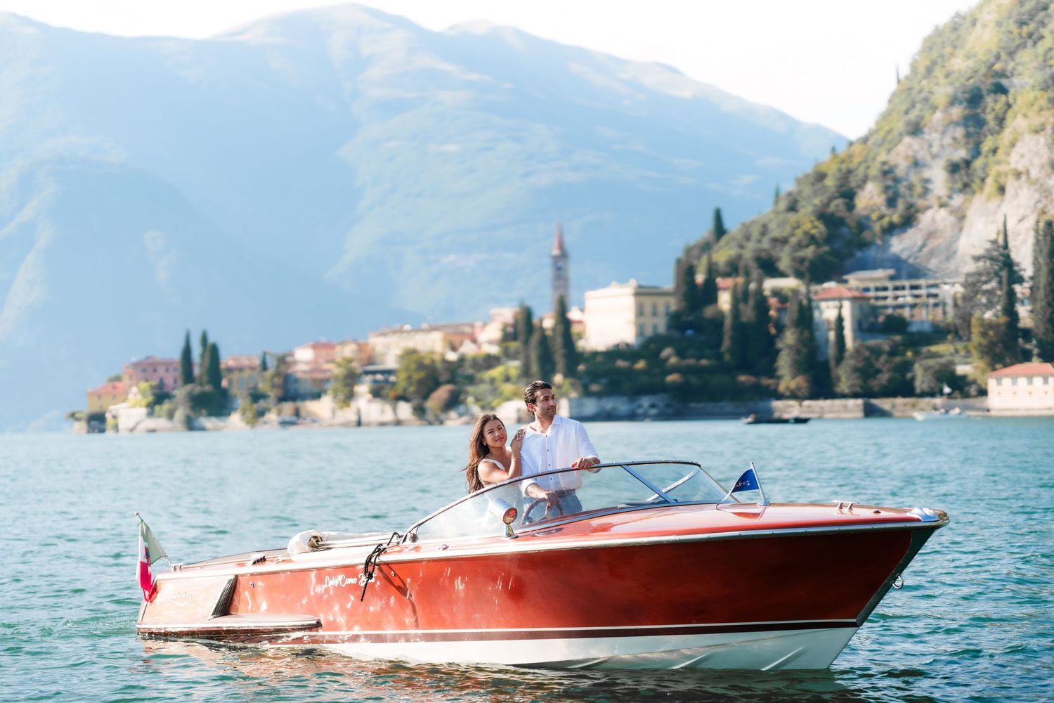 Couple riding a classic red speedboat on Lake Como with scenic Italian village and mountains in background.
