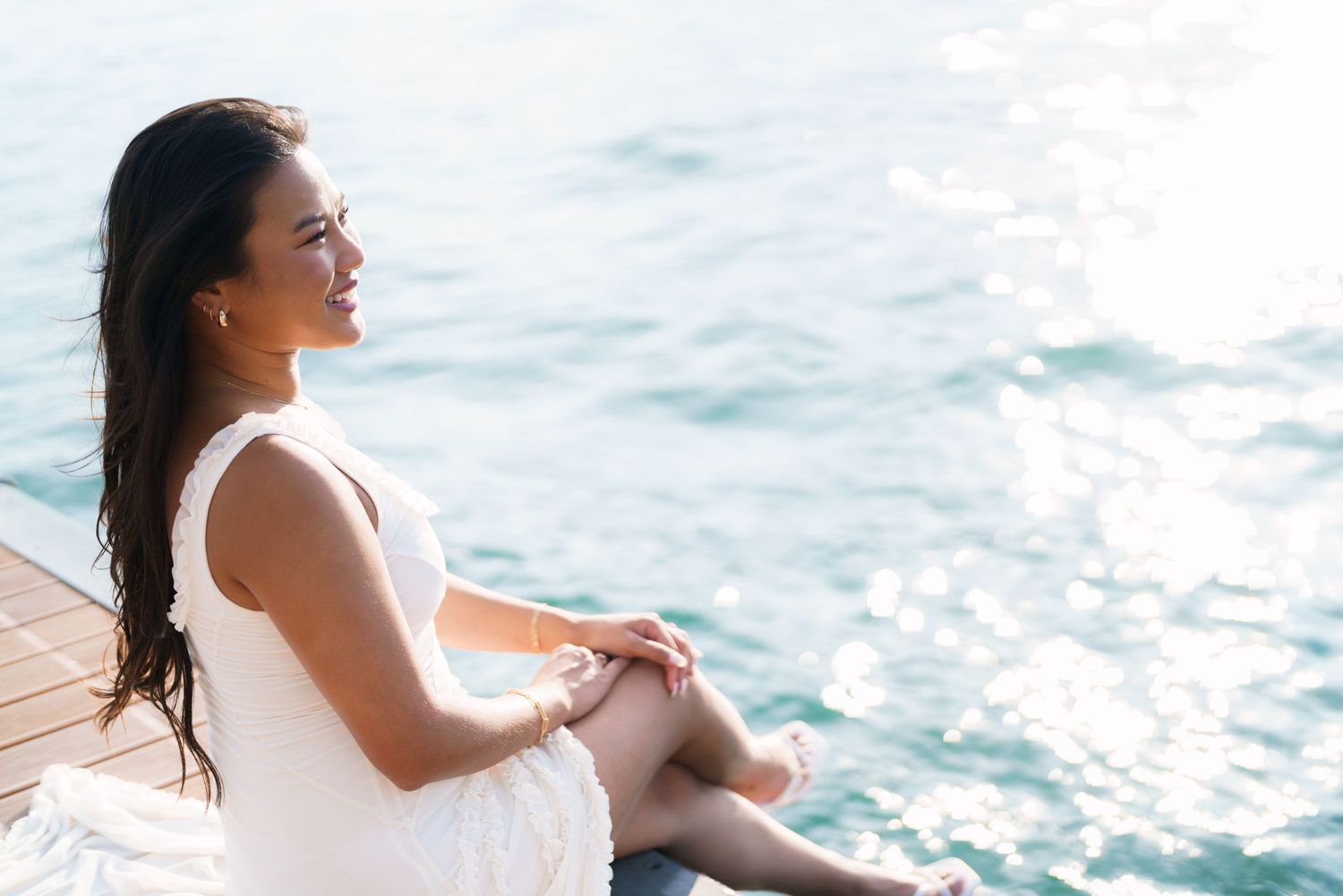 Young woman in white dress sitting on a wooden dock by the ocean, smiling peacefully in sunlight.