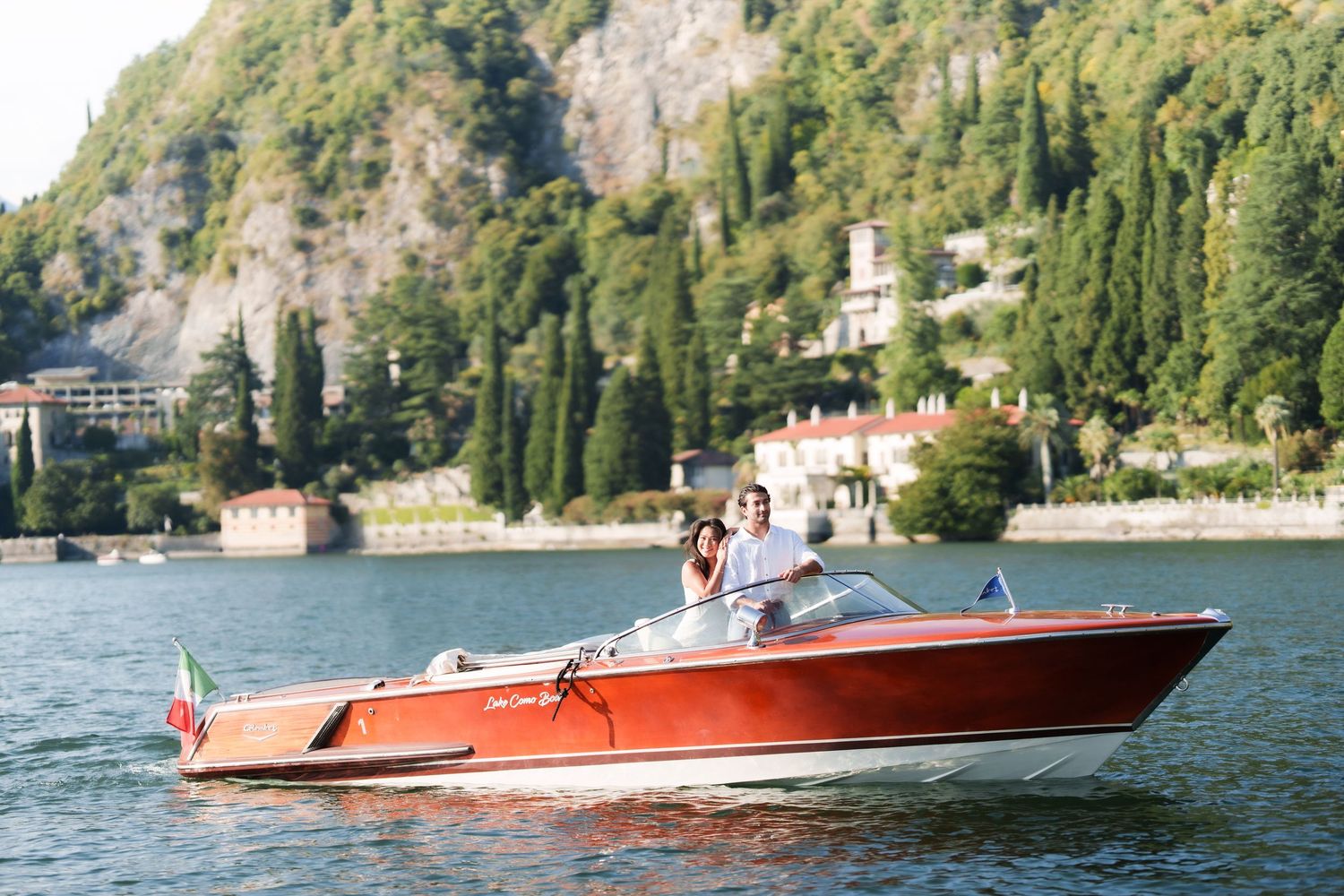 Couple riding a classic wooden Riva speedboat on Lake Como, Italy, with lush green hills and villas in the background.