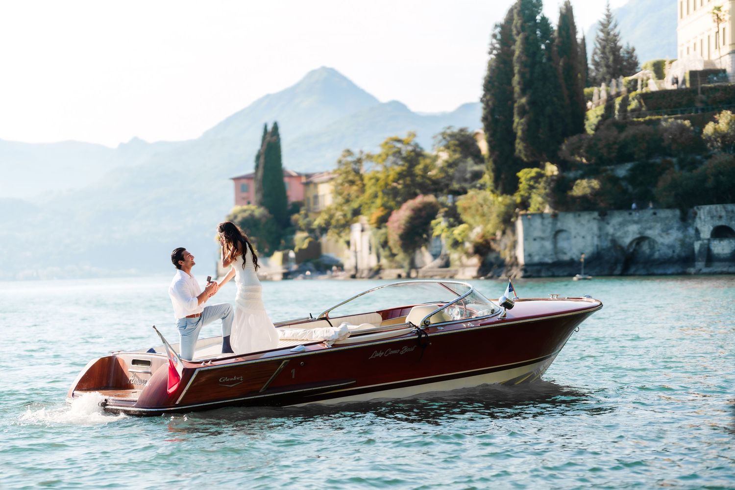 Couple embracing on a classic wooden boat on Lake Como with lush mountains and historic villa in the background.