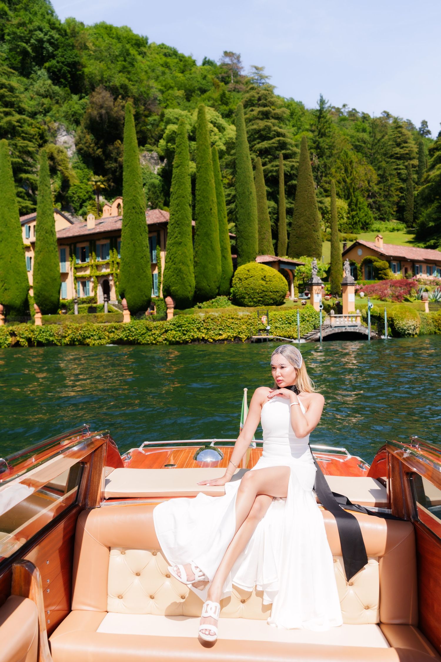 Elegant bride in white dress sitting on a luxury wooden boat on Lake Como with lush gardens and Italian villa in background.
