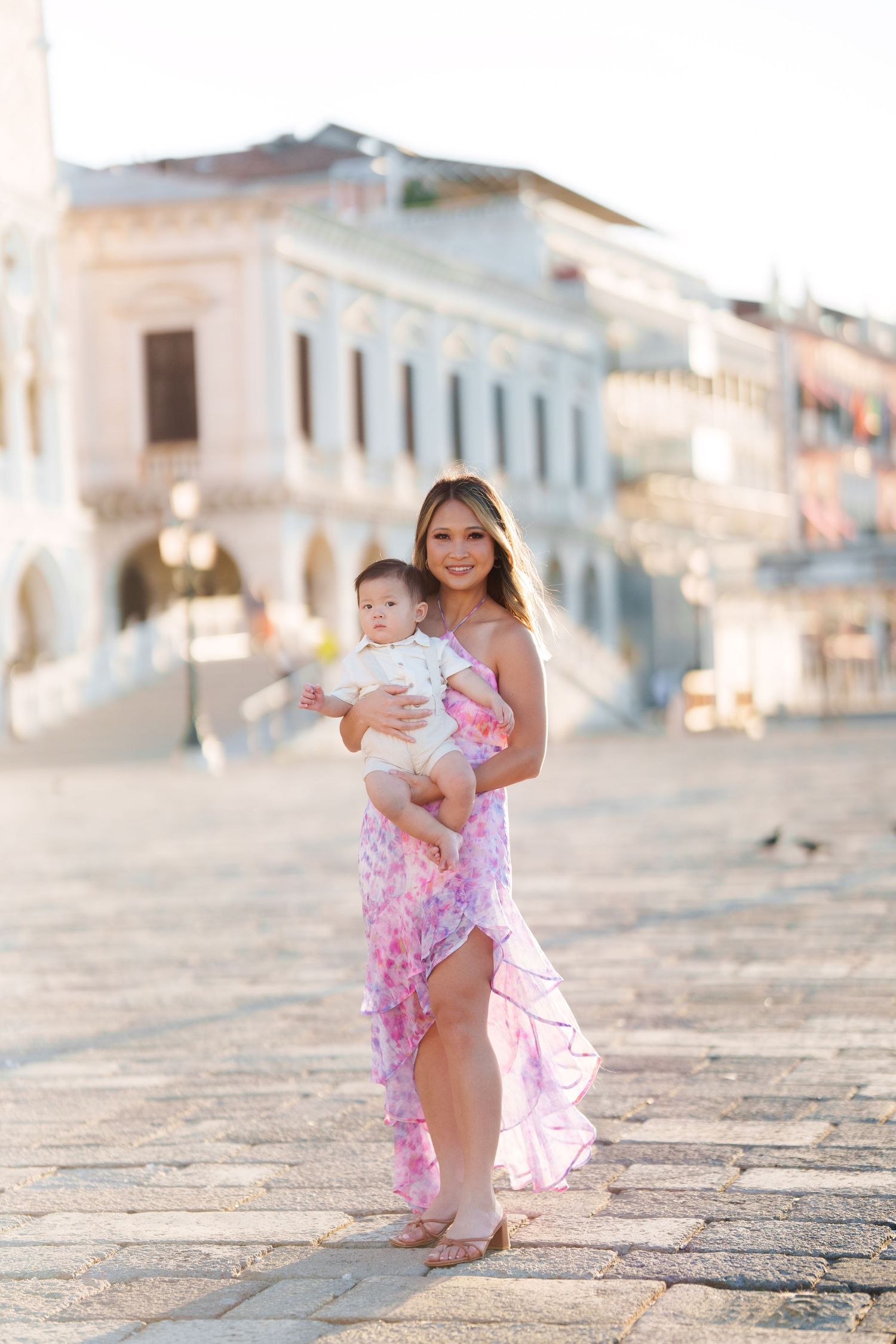 A smiling woman in a pink floral dress holds her baby on a sunlit cobblestone plaza with historic buildings behind her.