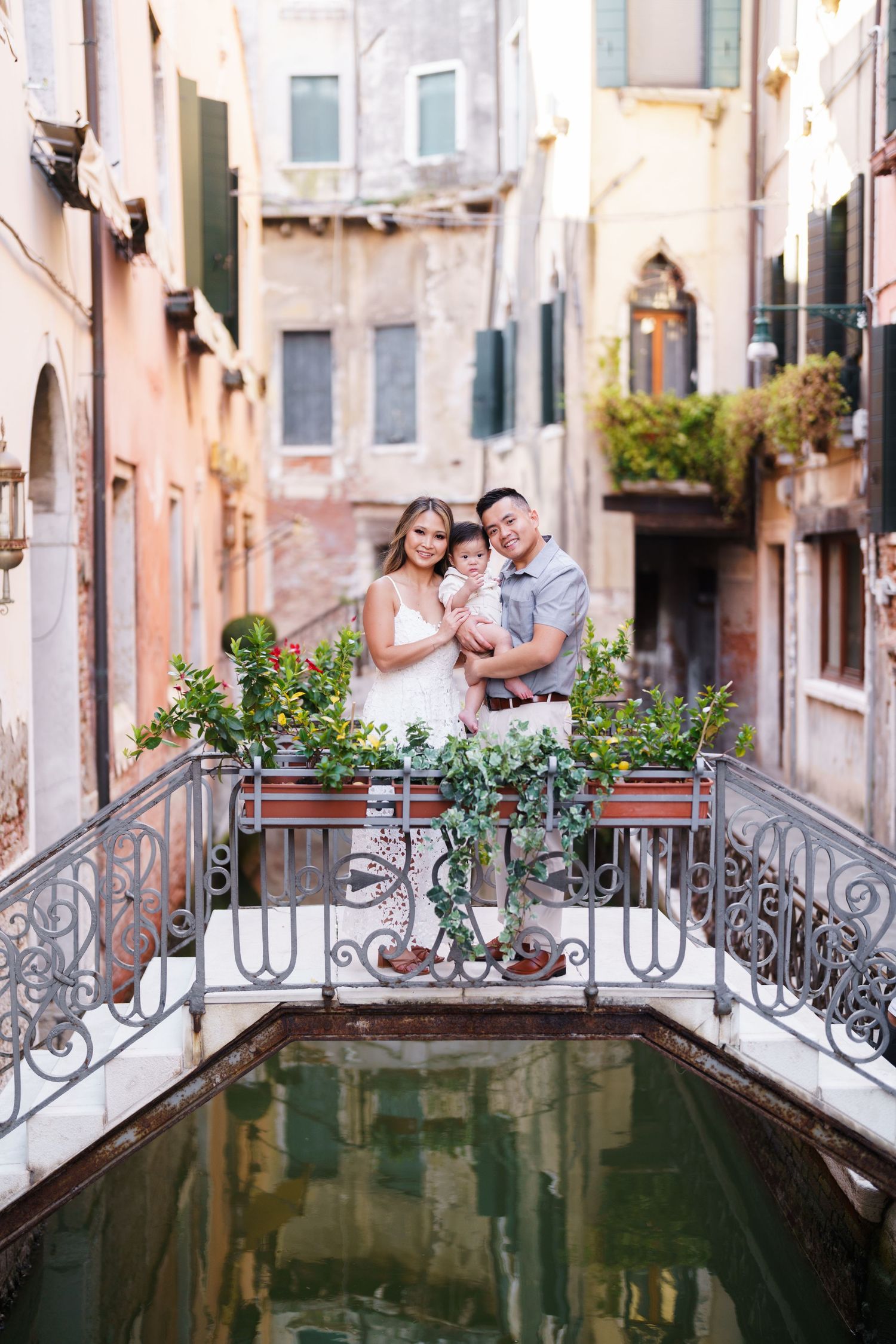 Family posing on ornate bridge over Venice canal, surrounded by lush greenery and historic buildings.