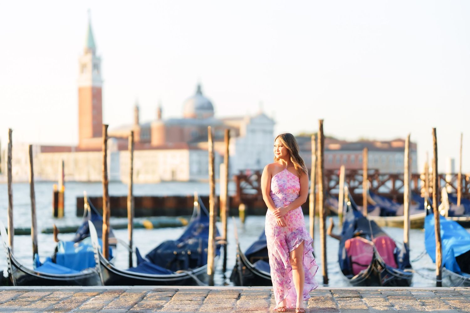 Woman in pink floral dress standing by Venice gondolas with San Giorgio Maggiore church in background.