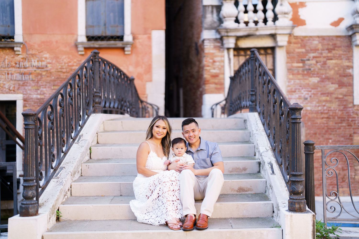 Happy family of three sitting on stone steps of a historic Venice building, parents smiling with baby in a sunny outdoor portrait.