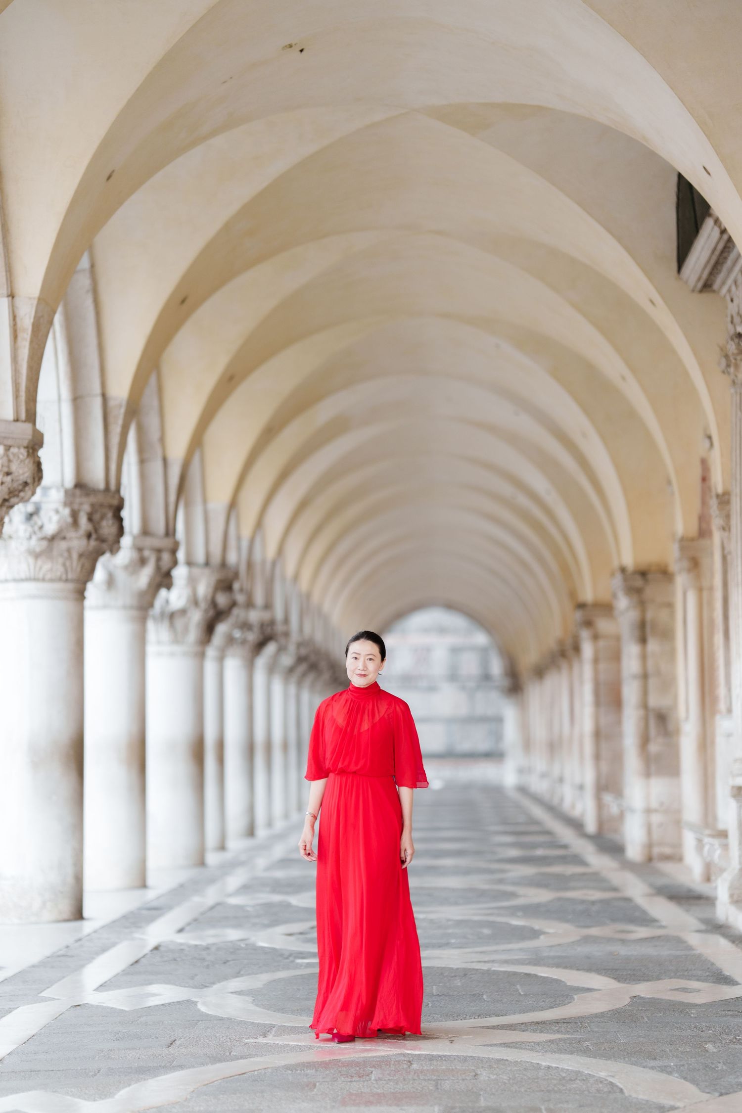 Woman in red gown standing under arched colonnade in Venice, Italy.