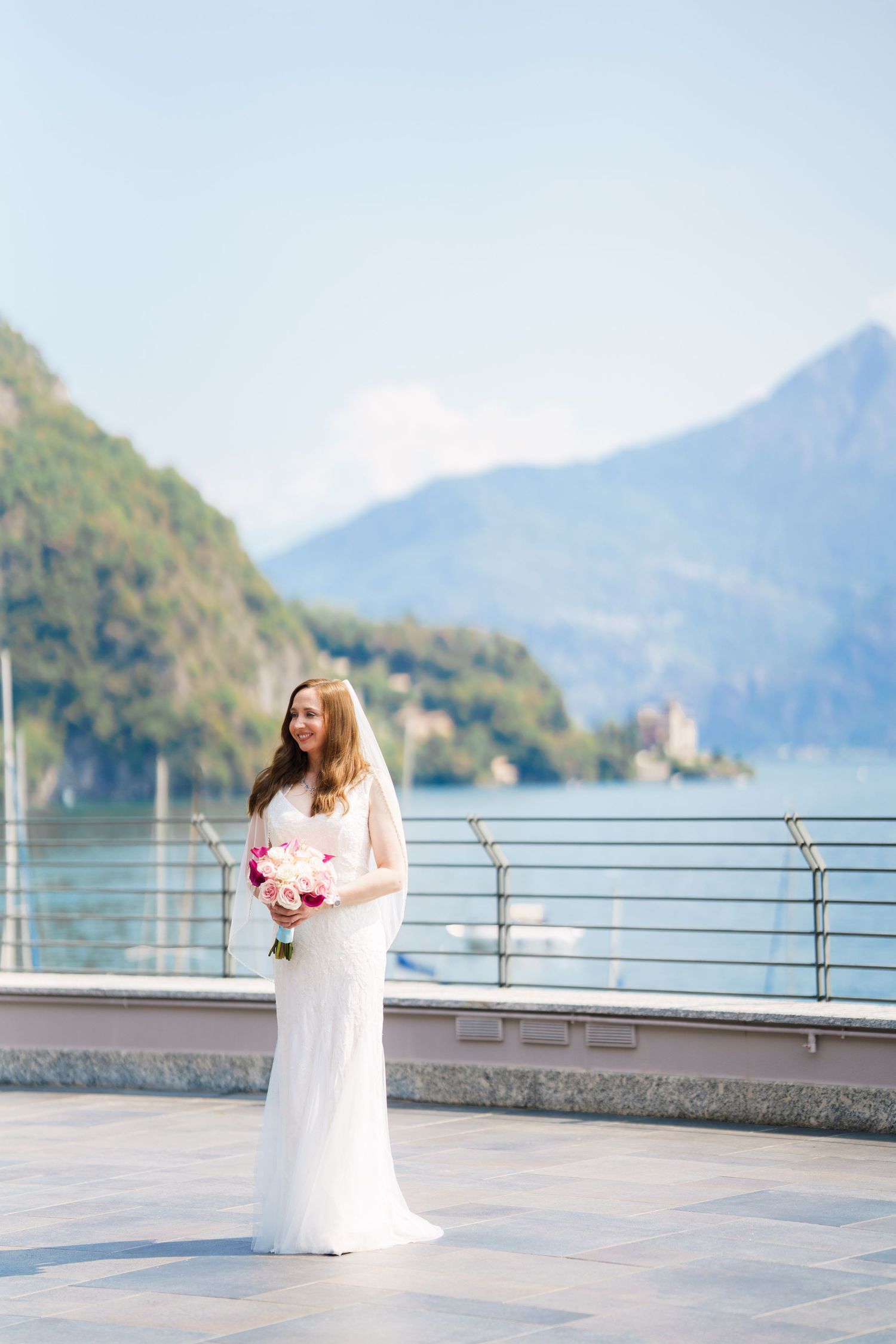 Bride in white gown holding pink bouquet on lakeside terrace with mountain backdrop on her wedding day.