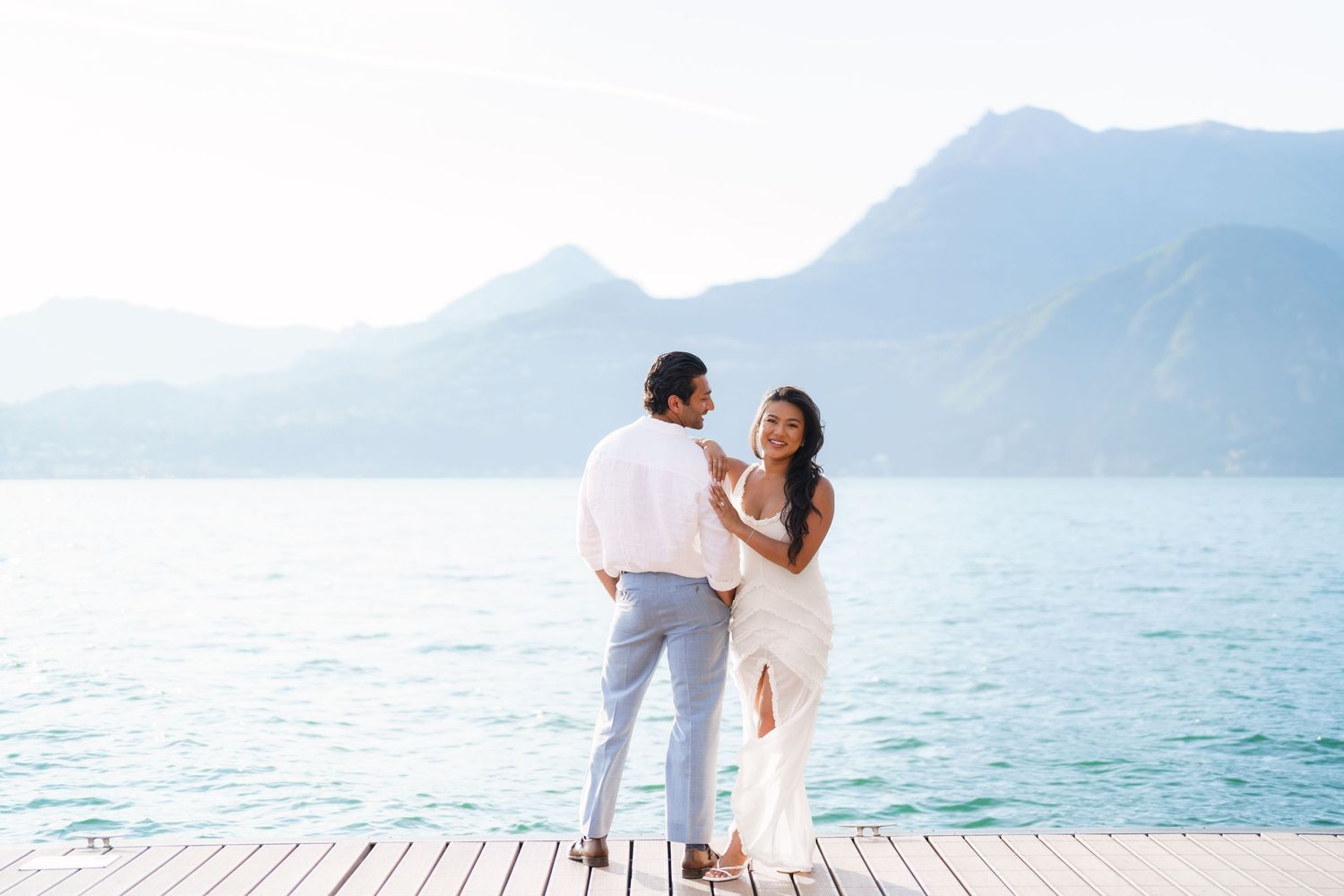 Couple in white attire sharing a romantic moment on a dock with turquoise lake and mountains behind.