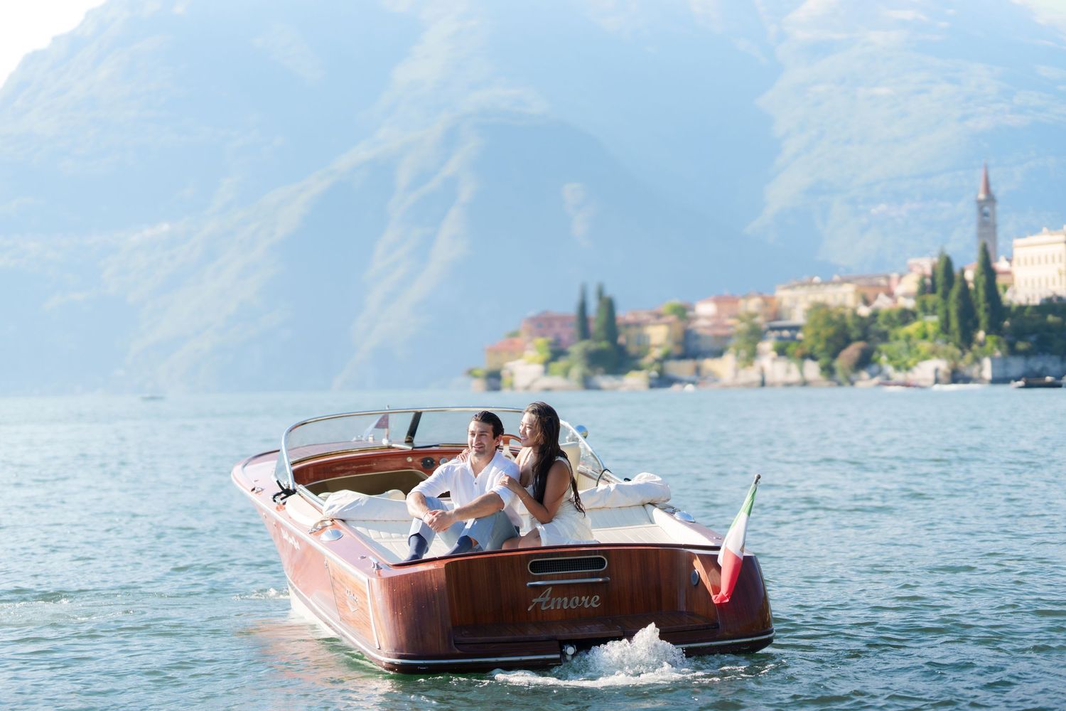 Couple enjoying a romantic boat ride on a serene lake with mountain backdrop and Italian village in distance.