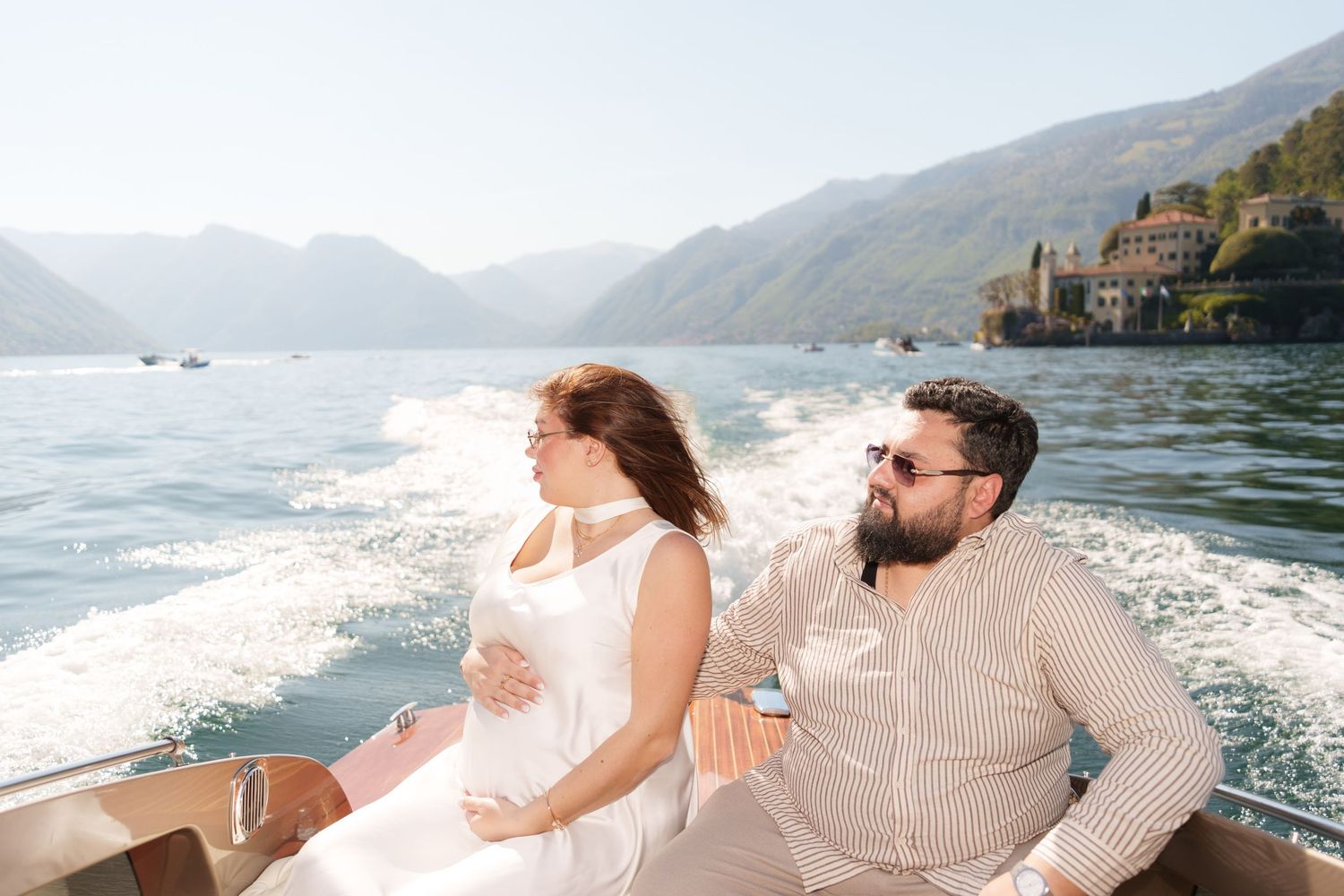 Pregnant couple riding a speedboat on a scenic lake surrounded by mountains and a picturesque village.