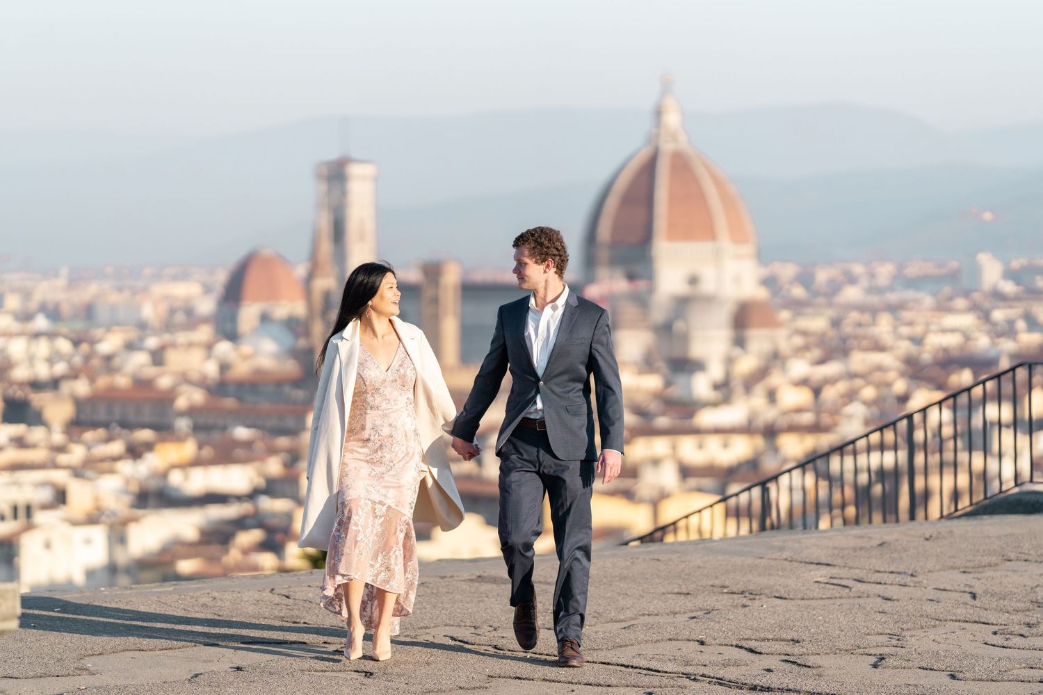 Couple in formal attire walking along a scenic overlook with Florence's Duomo cathedral visible in the background.