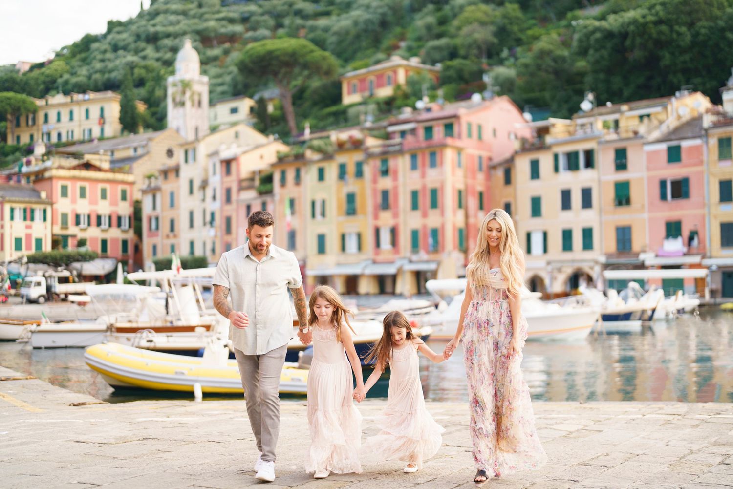 A family walks hand in hand along the colorful waterfront of Portofino, Italy with pastel buildings and boats in the harbor.