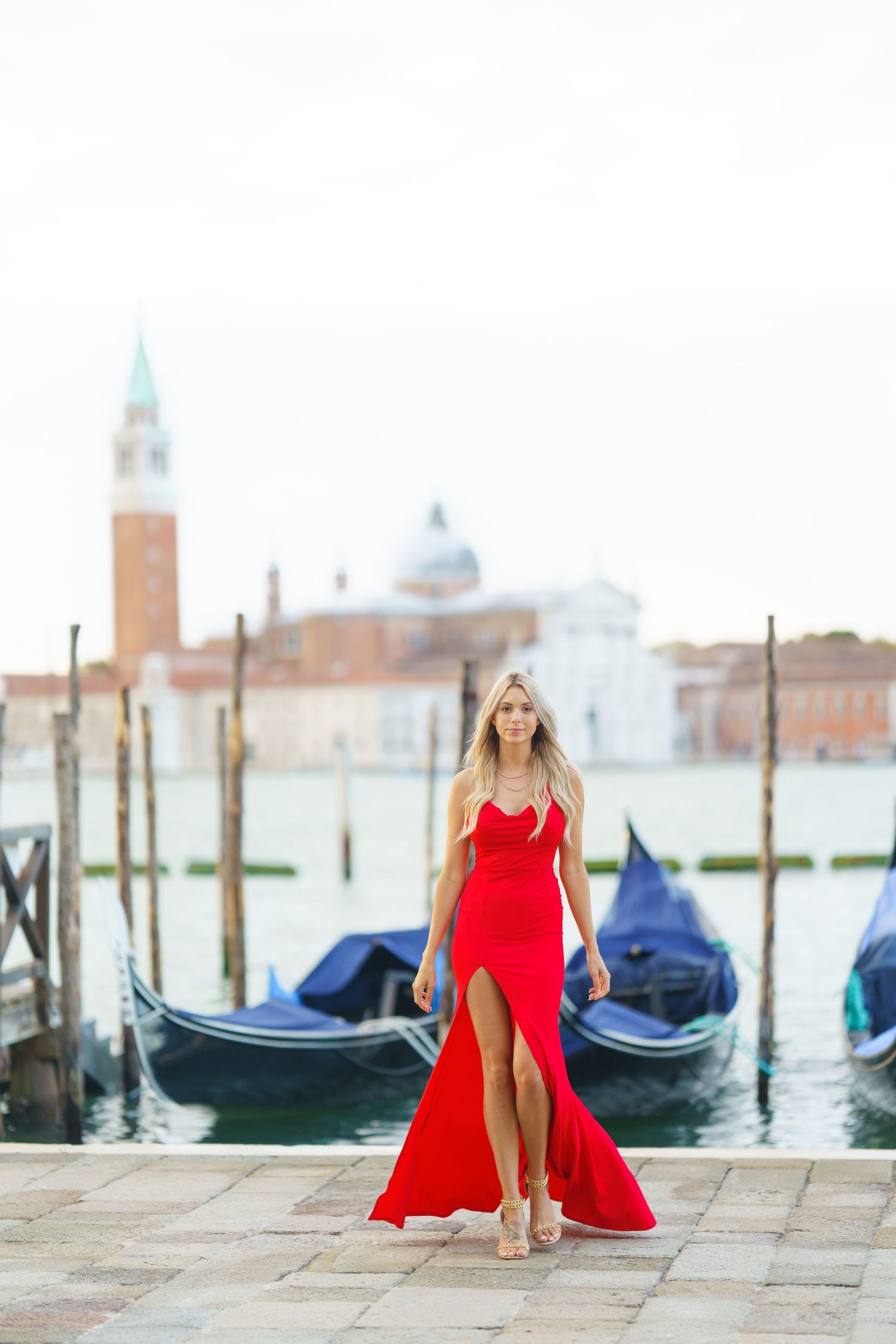 Woman in flowing red gown poses by gondolas in Venice with San Giorgio Maggiore church in background.