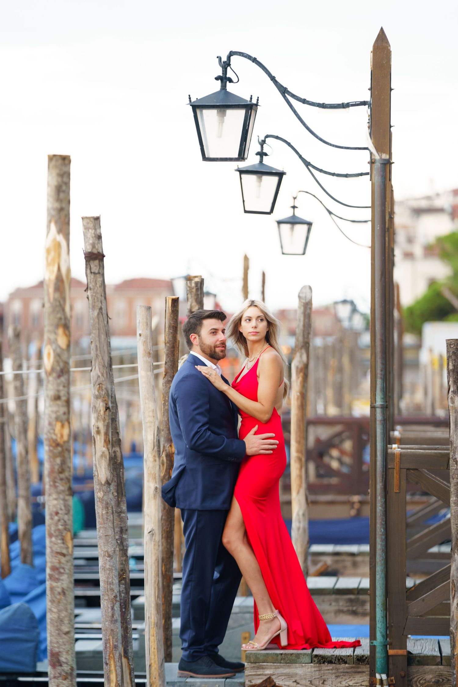 Romantic couple embraces on a dock with wooden posts and street lamps in Venice at sunset.