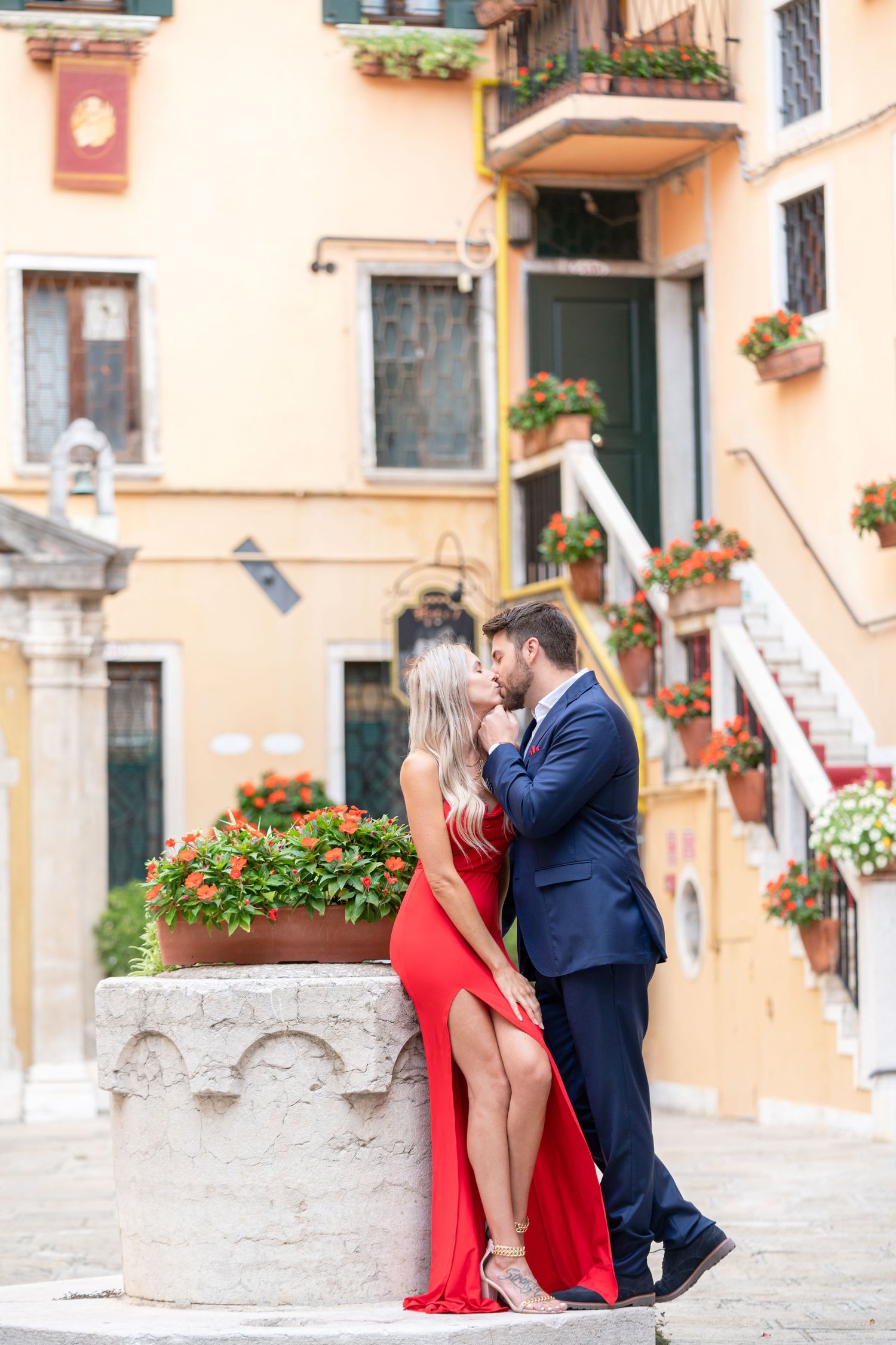 A romantic couple in formal attire embraces on stone steps against a peach-colored Mediterranean building with flowers.
