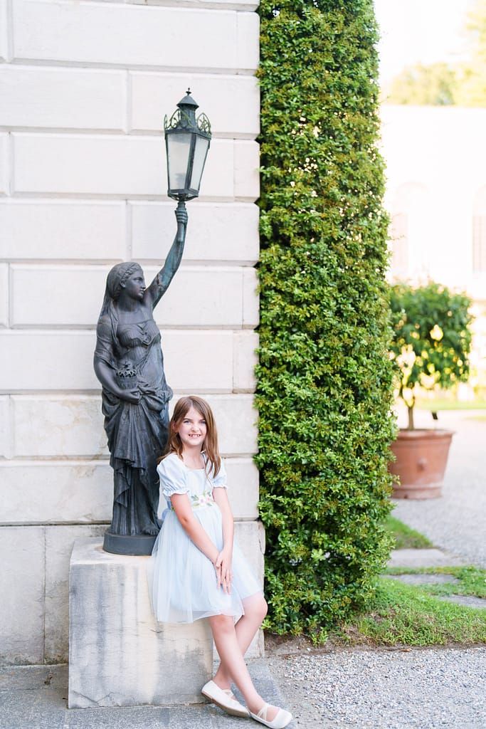 A decorative bronze statue holding a lamp post stands next to a tall hedge along a white building exterior.