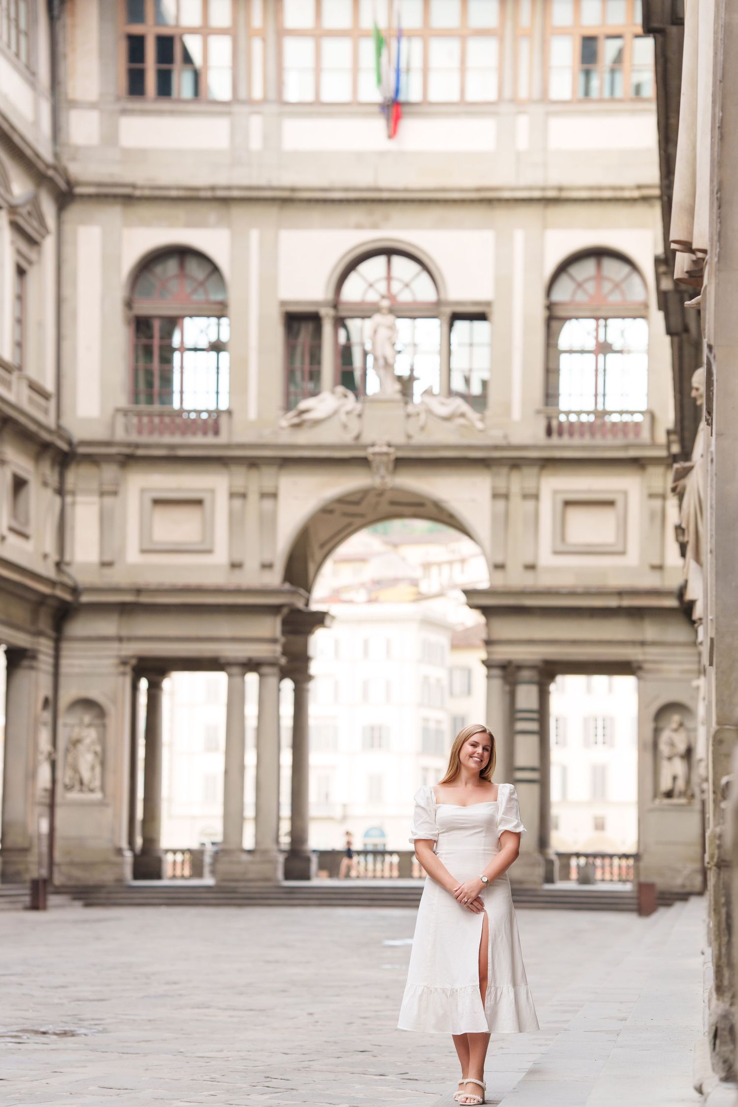 Woman in white dress poses in elegant gallery with ornate architecture and arched windows.