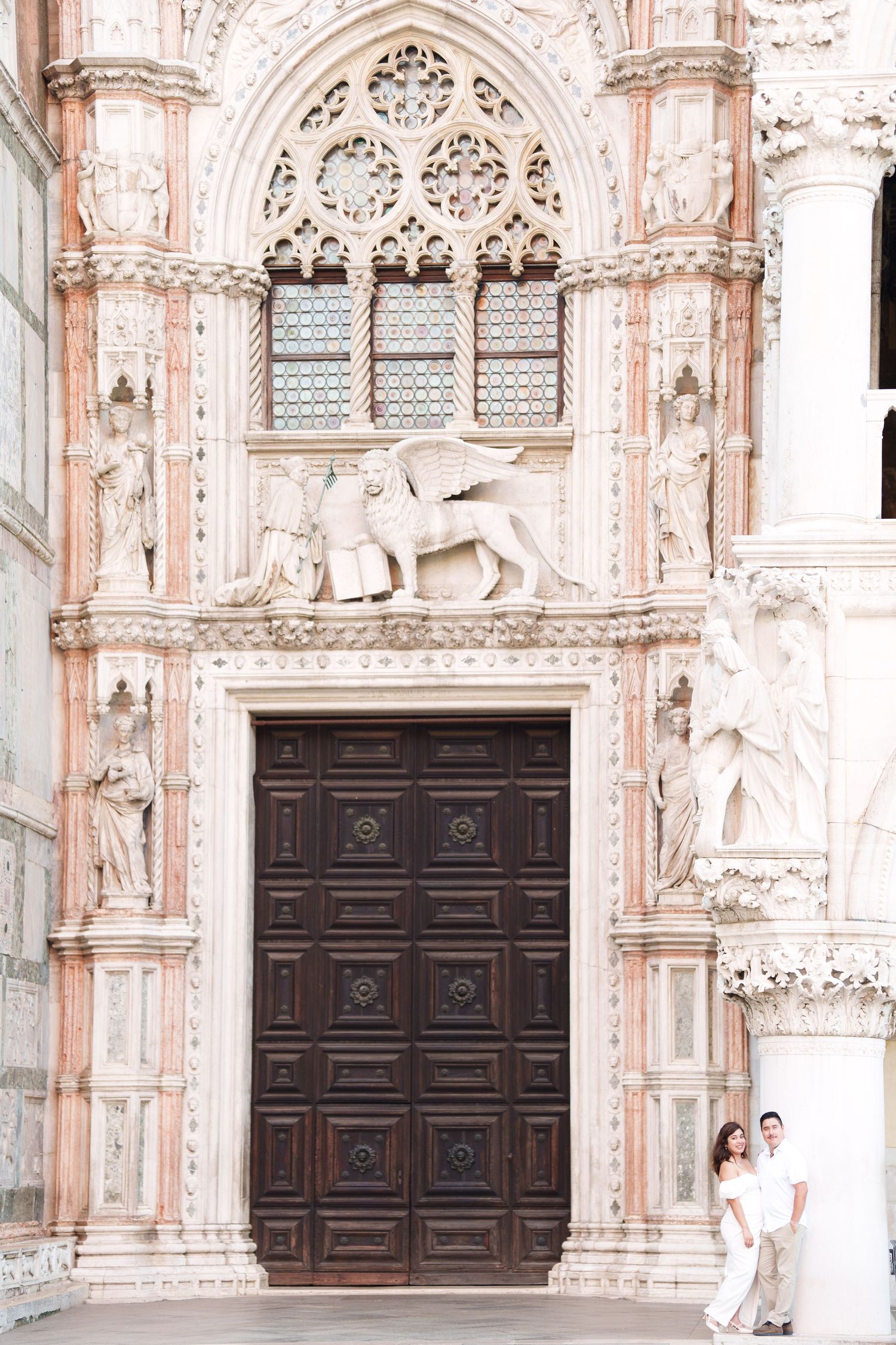Ornate Gothic cathedral entrance with detailed stone carvings and large wooden doors in a pink-tinted facade.