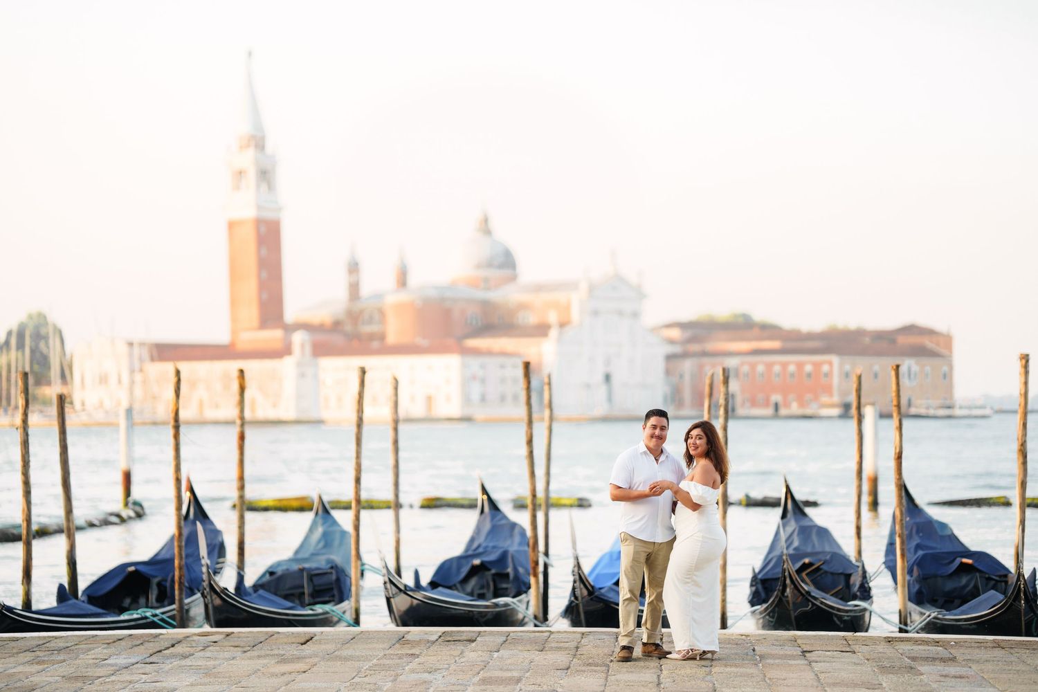 photographer veniceRomantic couple stands by traditional gondolas in Venice with San Giorgio Maggiore church in the background at sunset.